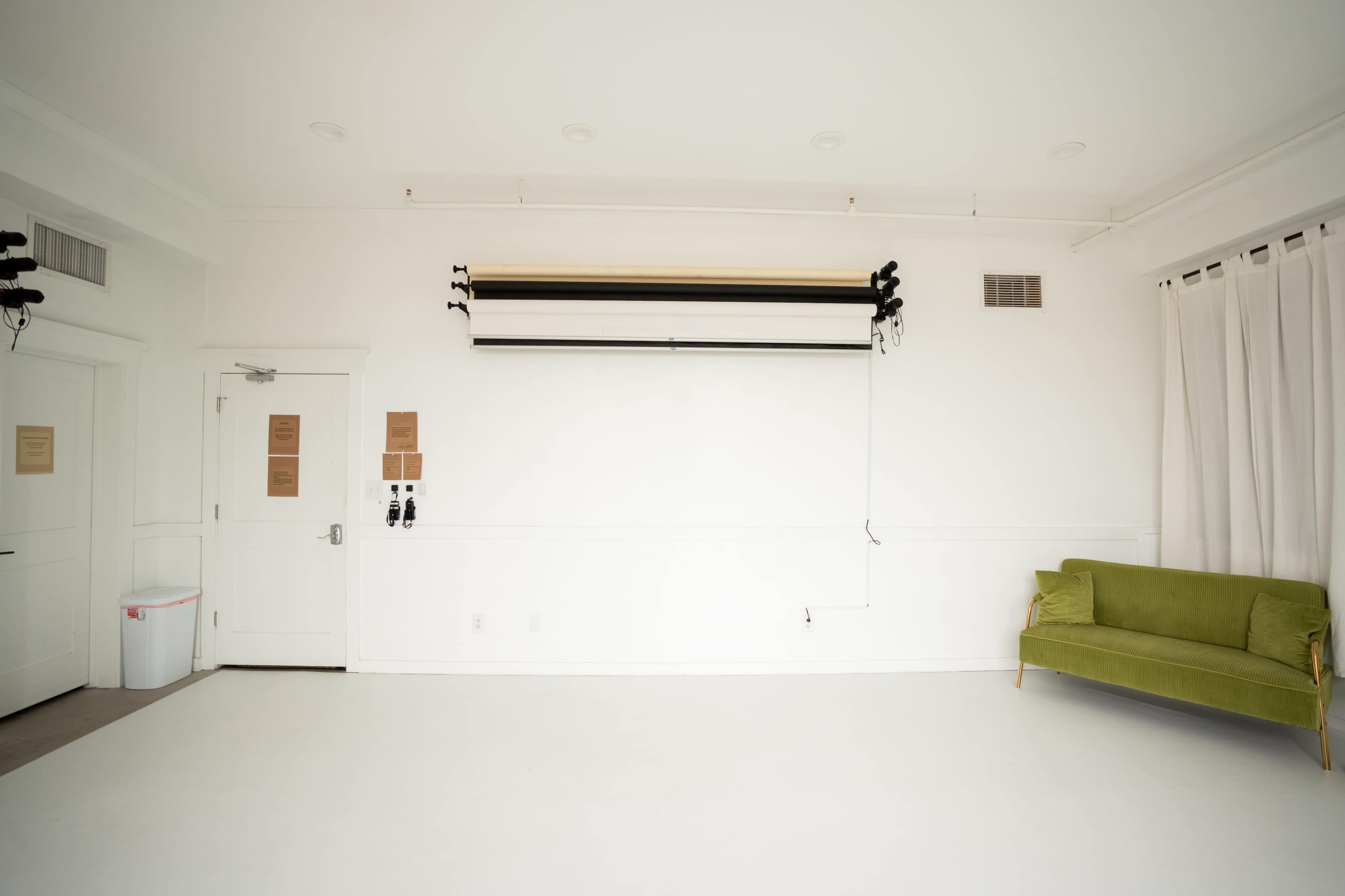 The image shows a bright, empty room with white walls and flooring, featuring a green sofa in the corner, a door on the left, and window treatments.