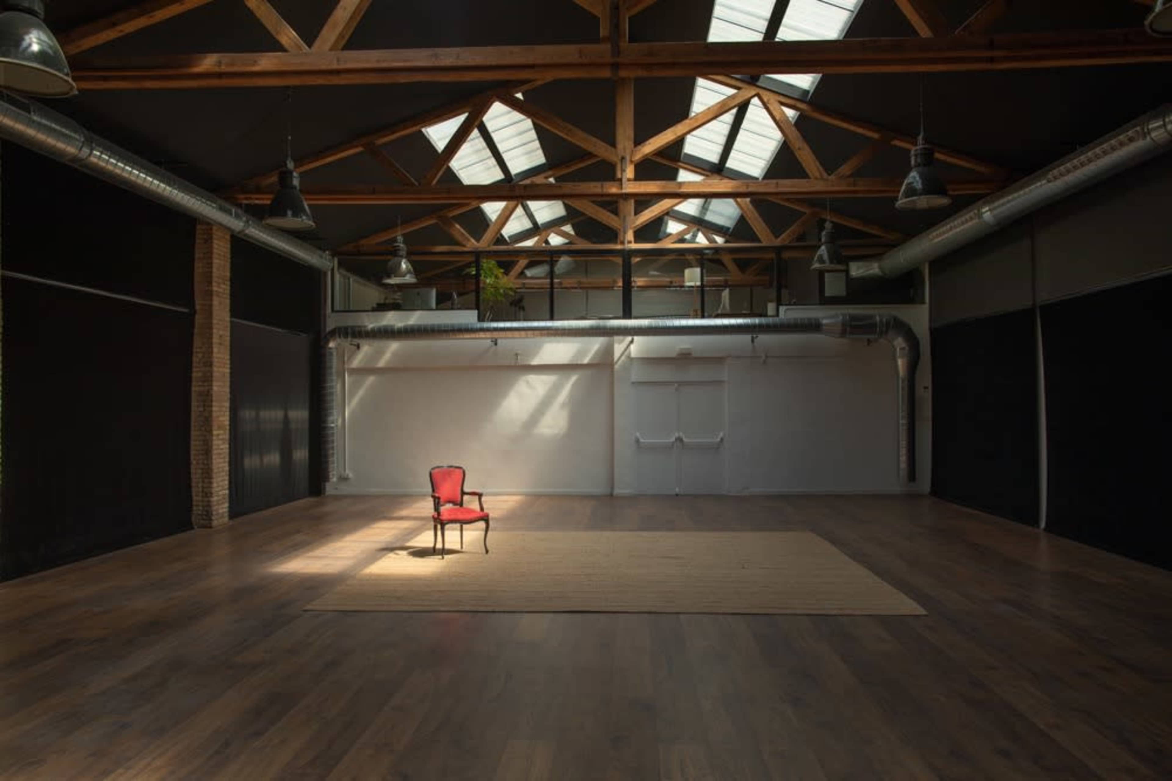 A spacious, empty room with wooden beams, a single red chair on a rug, and large windows allowing natural light to enter.