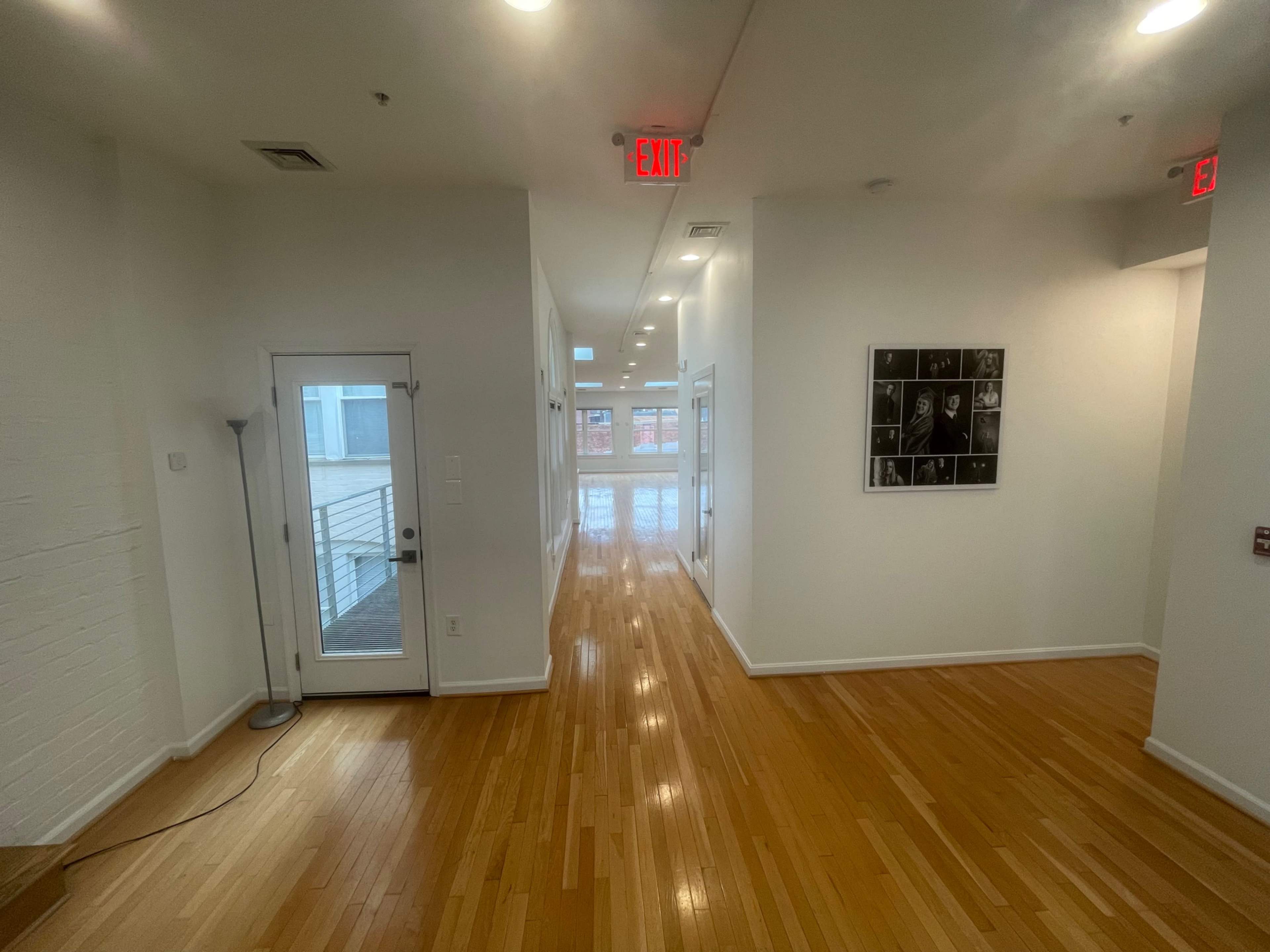 A well-lit corridor features a doorway on the left and a series of framed photographs on the right, with polished wooden flooring extending down the hall.