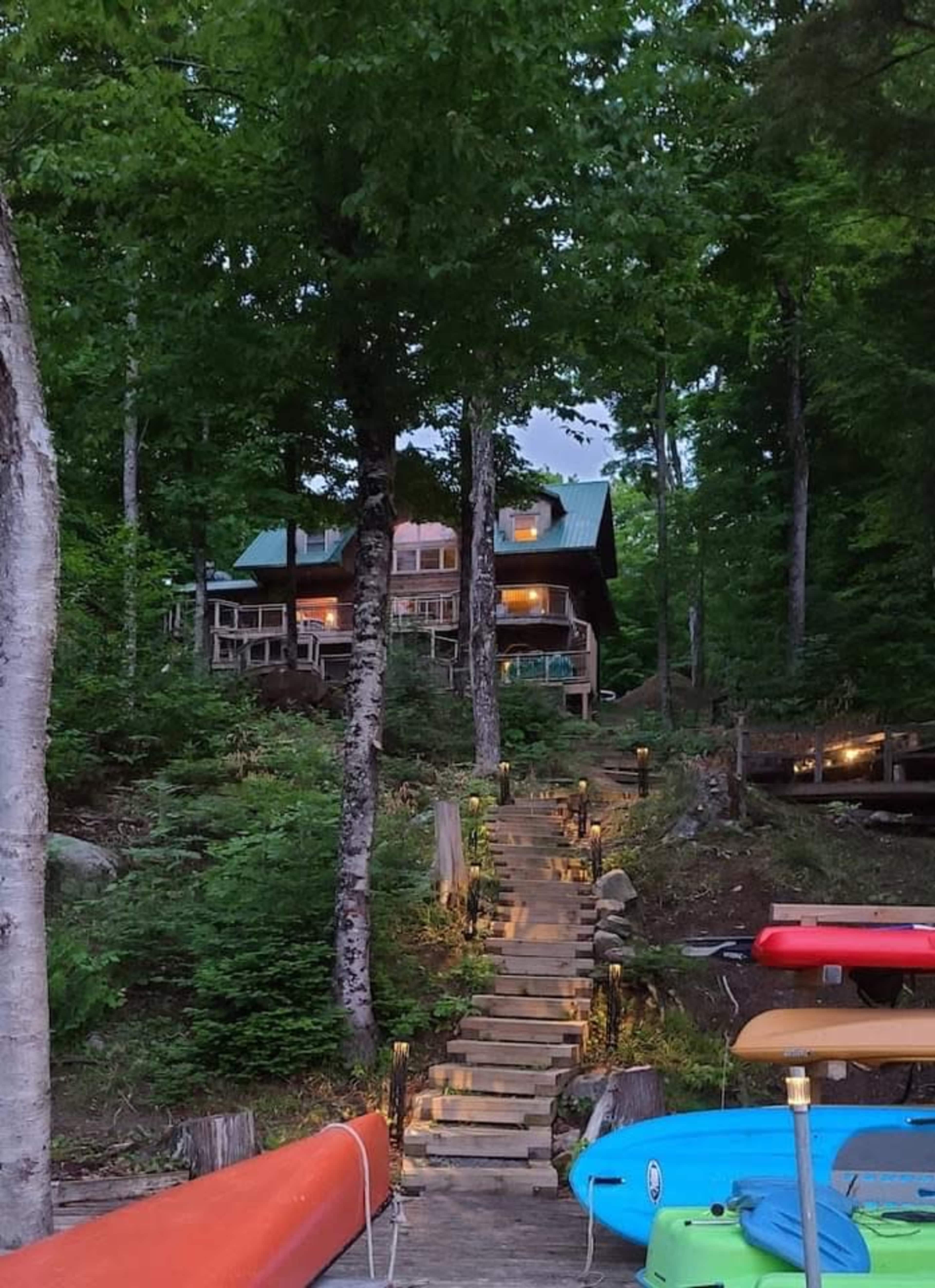 A wooden cabin sits atop a stone staircase surrounded by trees, with canoes lined at the water's edge.