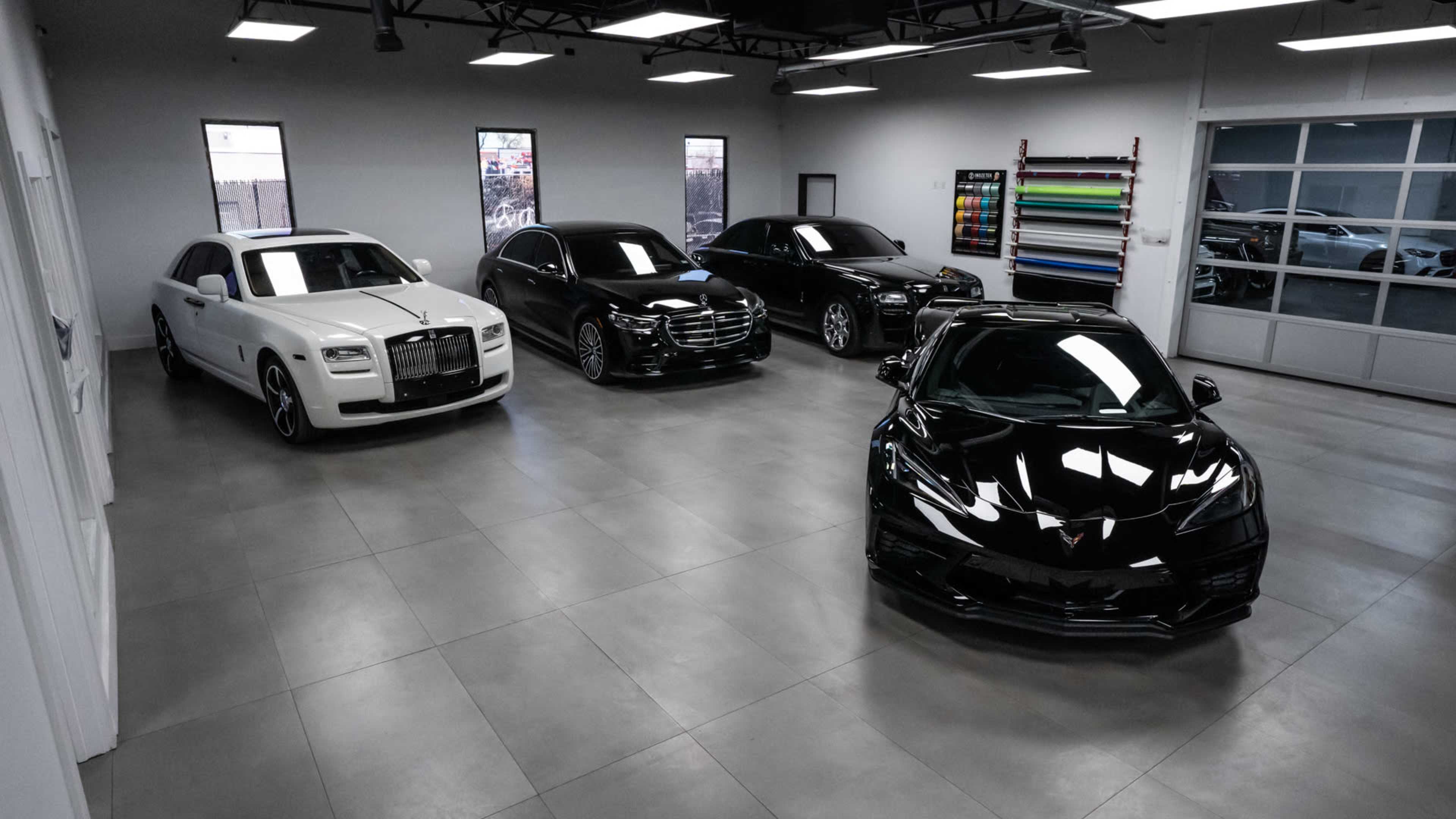 Four luxury cars, including a white Rolls Royce, a black Mercedes-Benz, a black sedan, and a black Corvette, are parked in a well-lit garage with gray flooring.