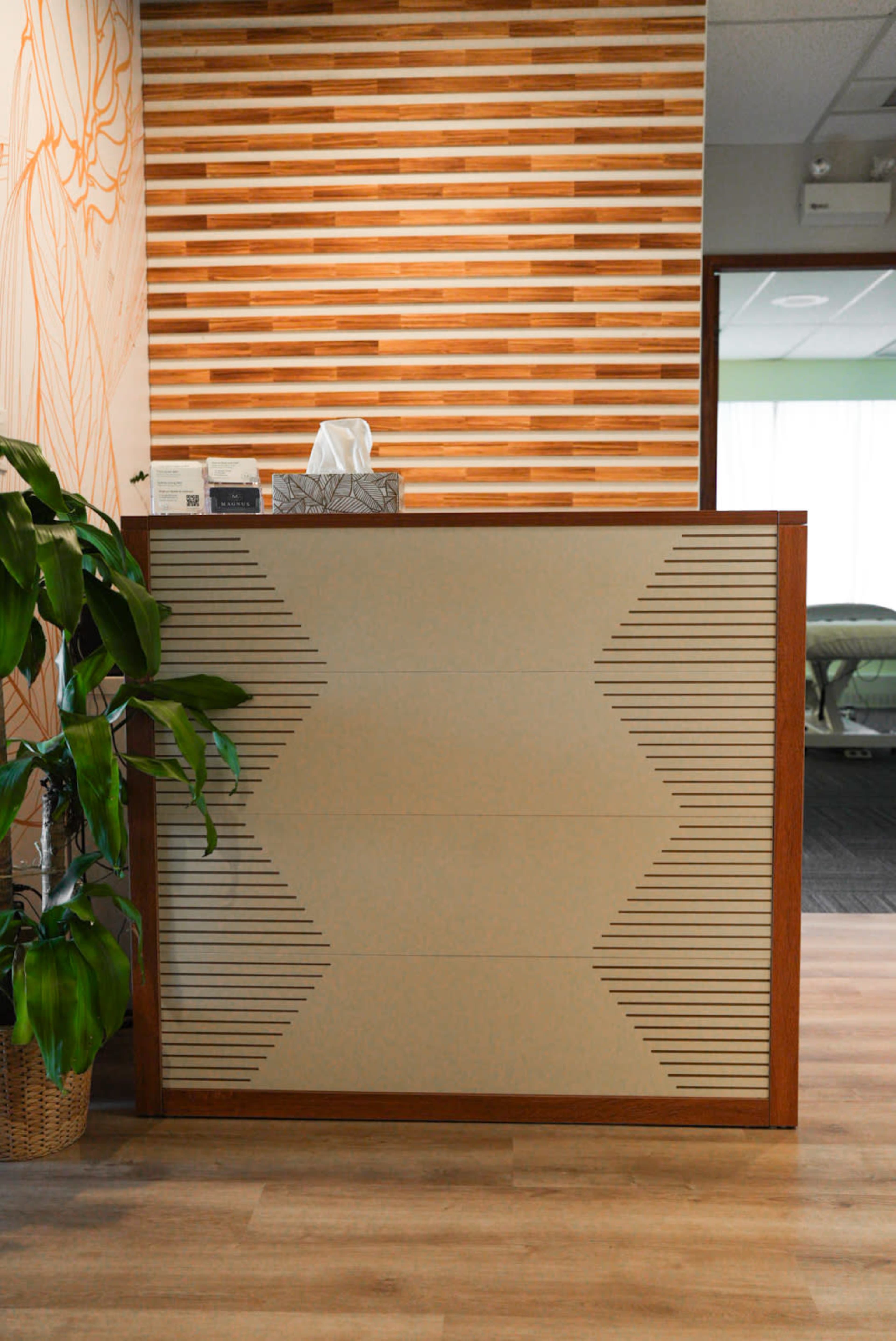 The image shows a reception desk with a decorative panel and a plant beside it, against a backdrop of a wooden accent wall.