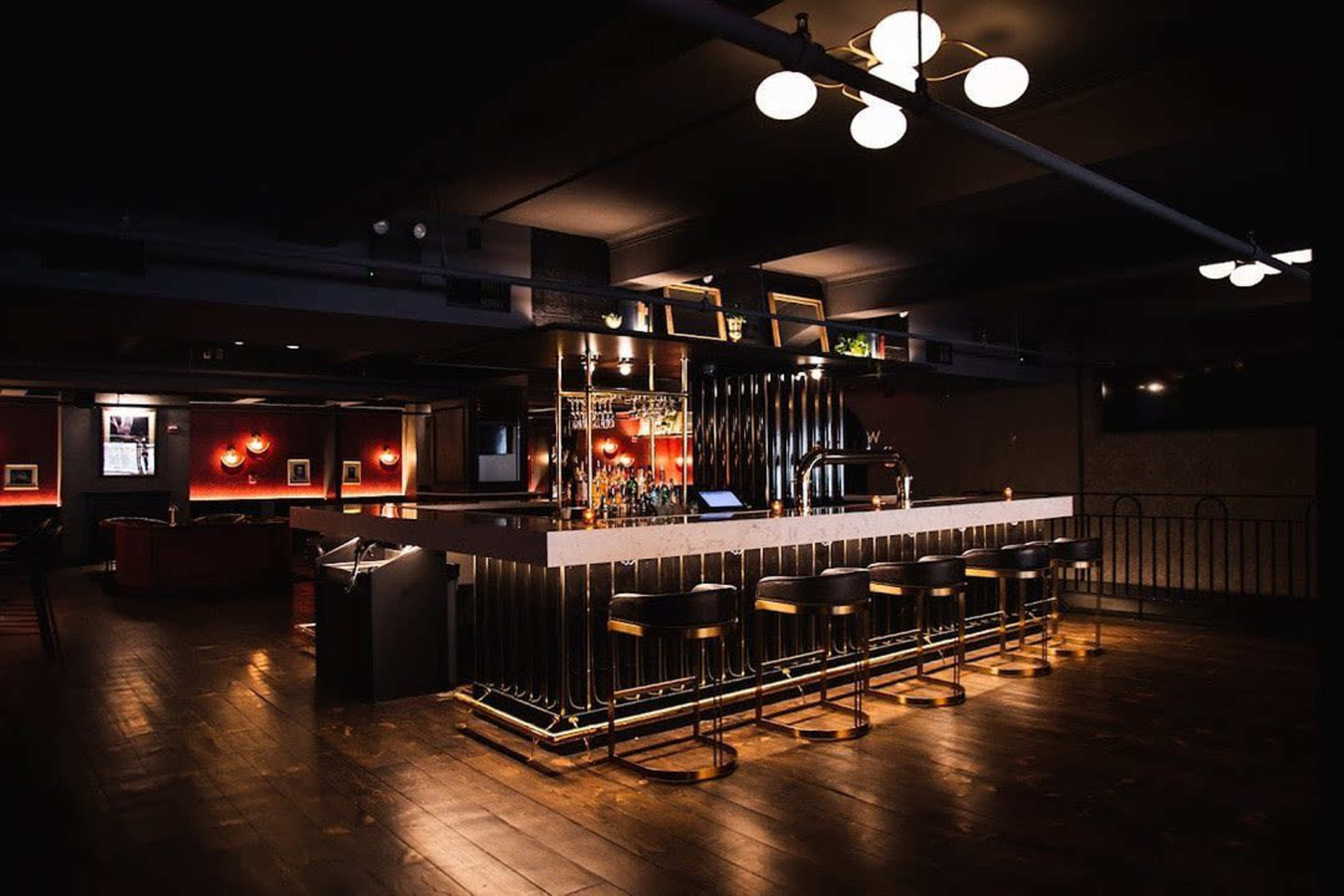 A dimly lit bar features a sleek, modern counter with high stools and a well-stocked liquor shelf in the background.