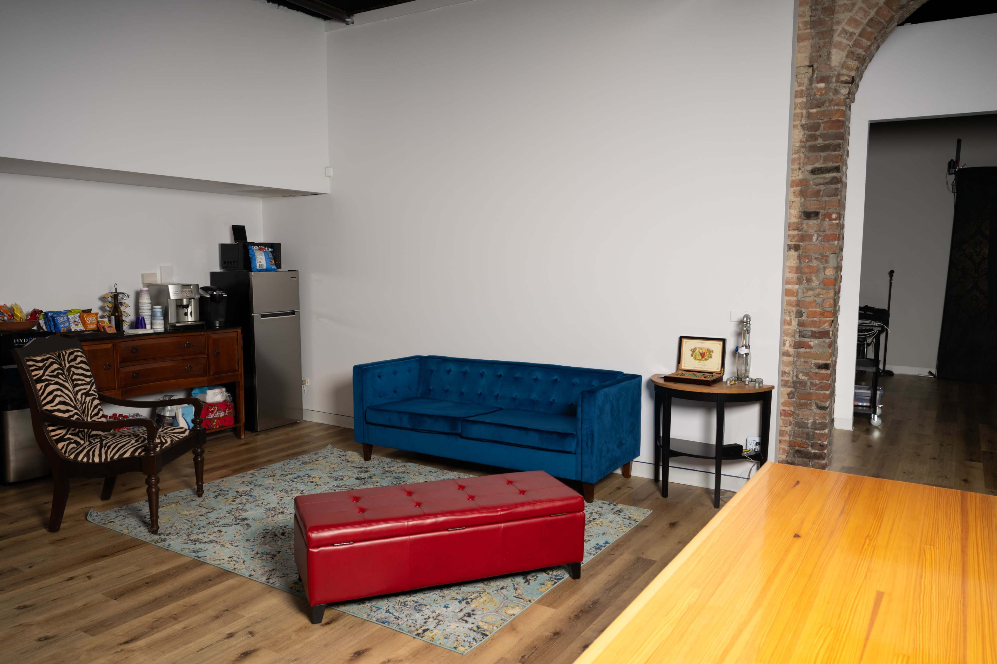 A modern room with a blue tufted sofa, a red ottoman, a wooden chair with a patterned upholstery, and a small table, all set against a backdrop of a brick wall.