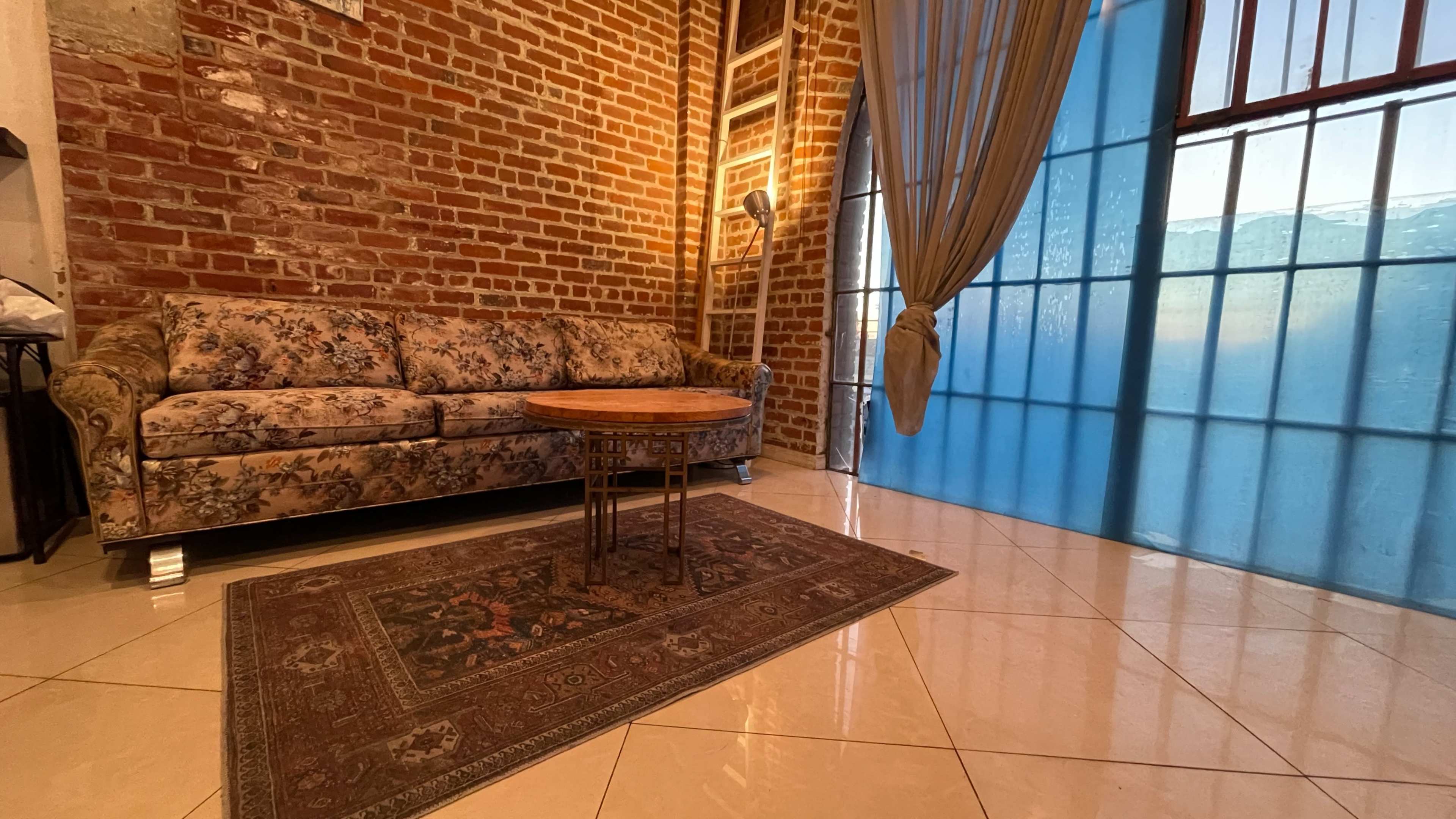 The image shows a cozy living area with a floral-patterned sofa, a round wooden coffee table, and a large window with sheer curtains, all set against a backdrop of exposed brick walls and polished tile flooring.