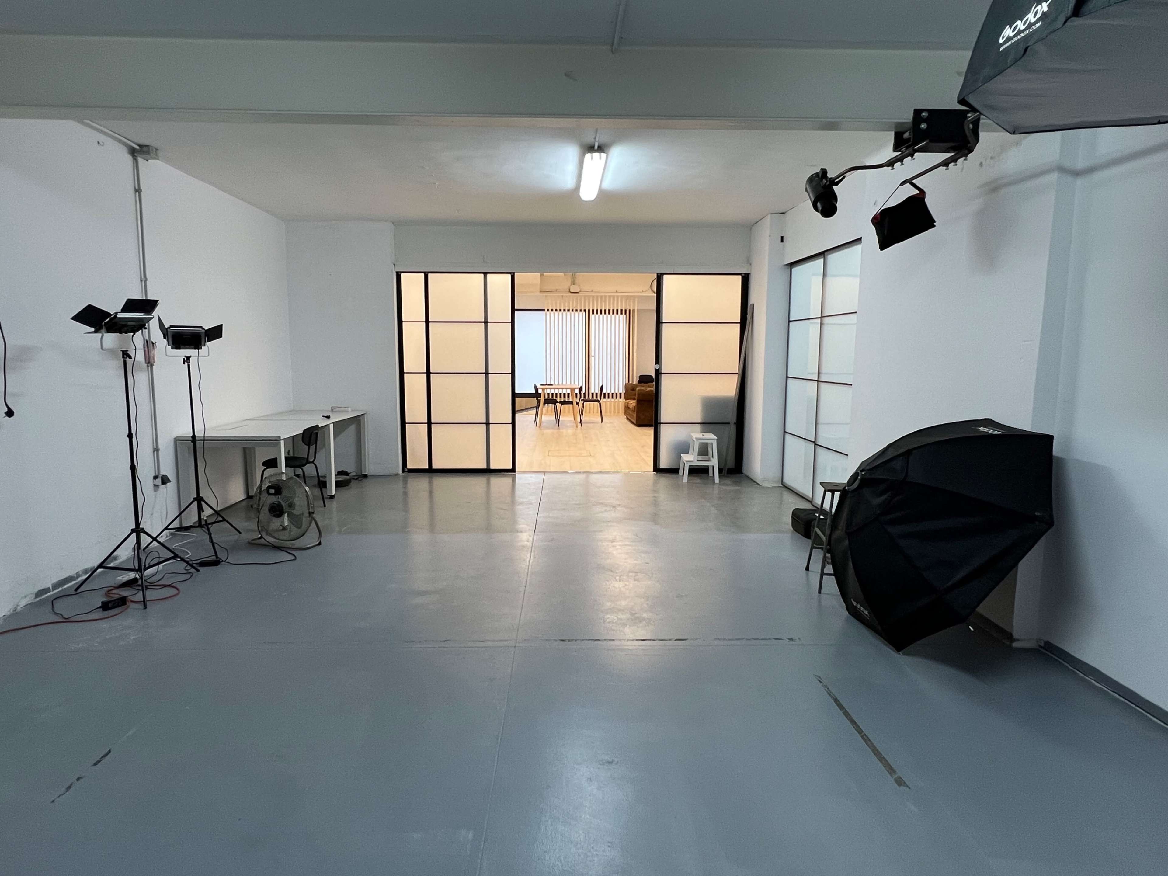 The image shows a spacious studio space with studio lights, a fan, and a softbox on the left, leading to a room with a table and chairs in the background.