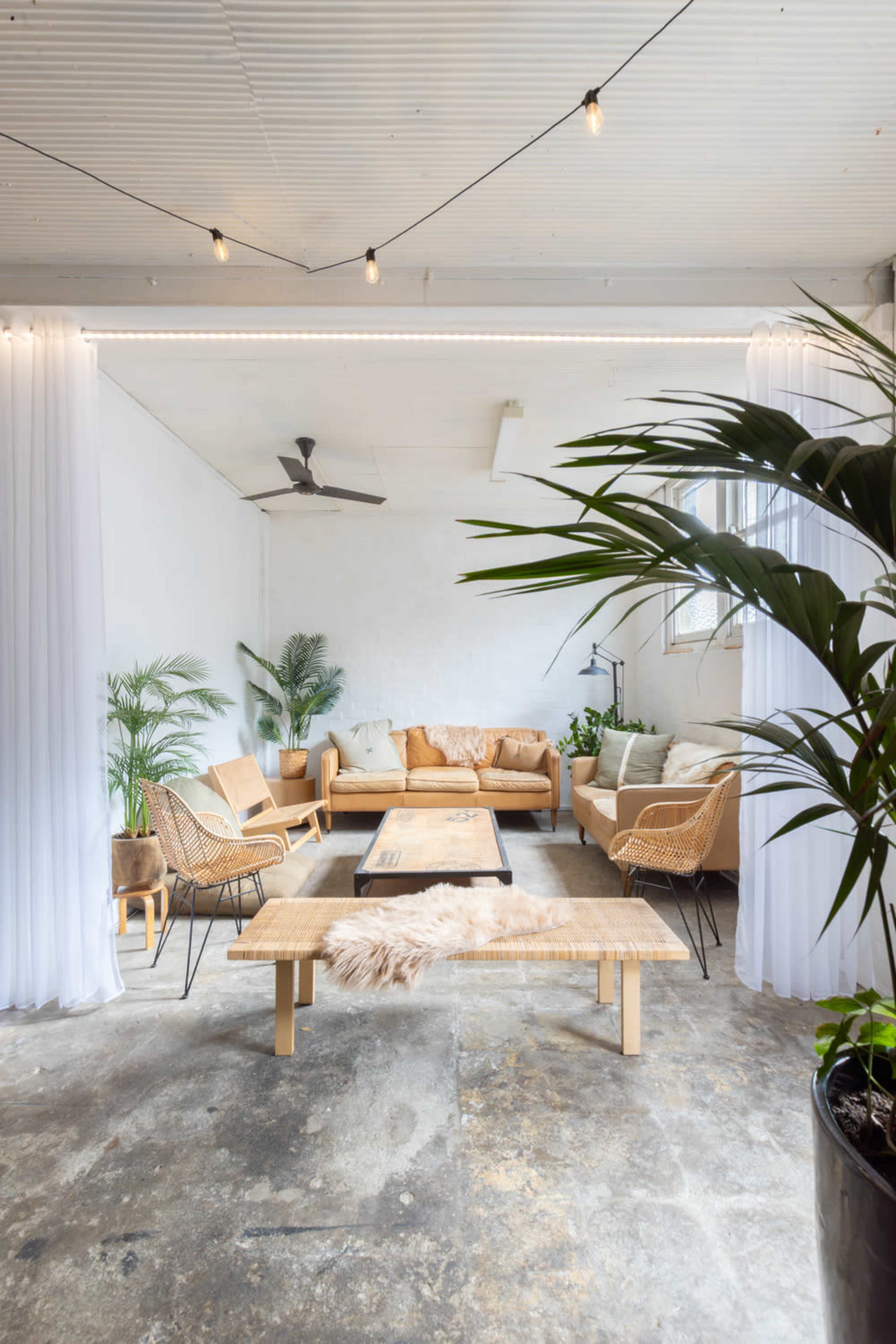 The image shows a spacious, brightly lit lounge area with a mix of beige furniture, wooden accents, and potted plants, separated by sheer white curtains.