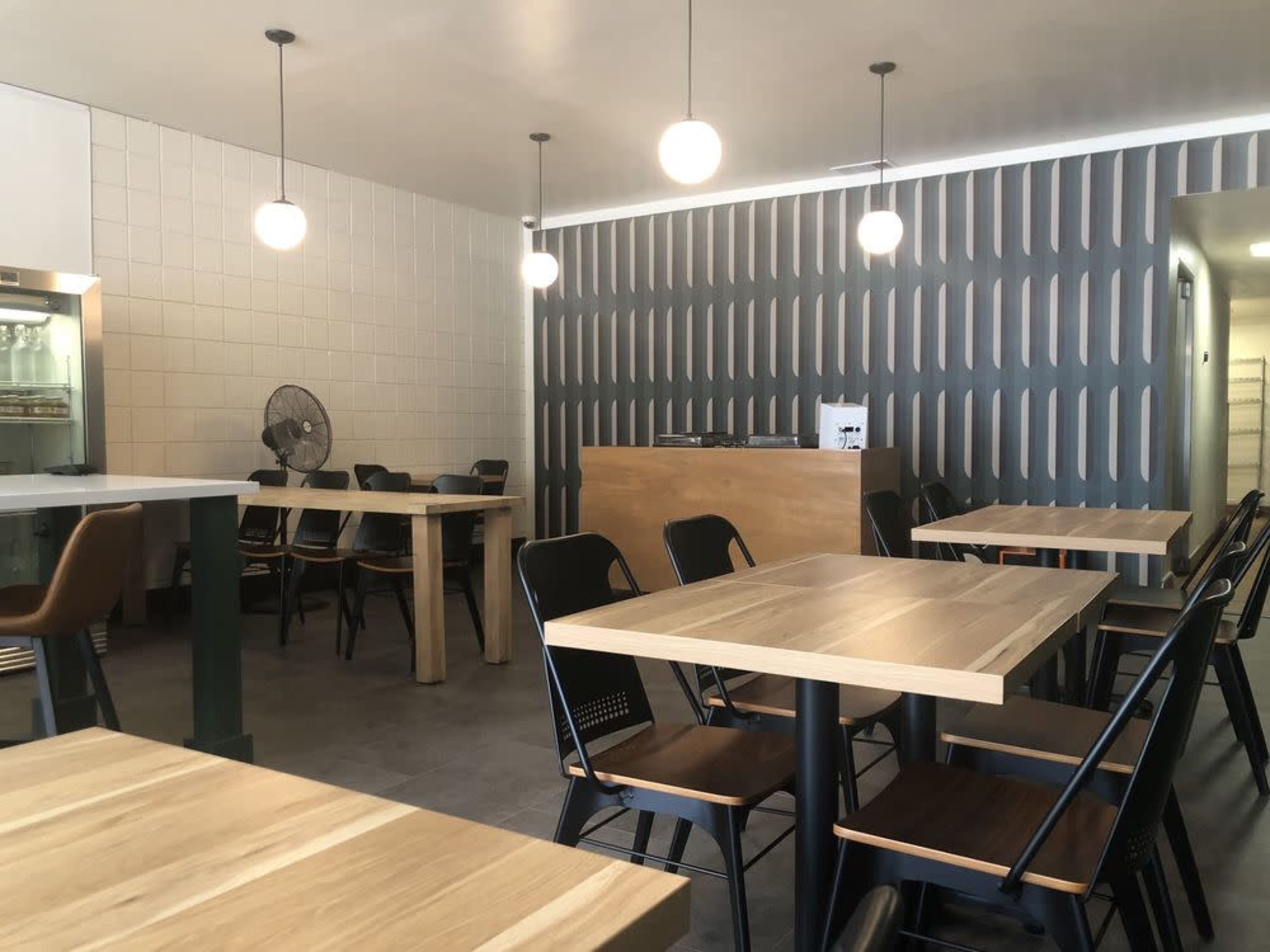 The image shows a modern café interior featuring wooden tables and black chairs, with a decorative wall and overhead lighting.