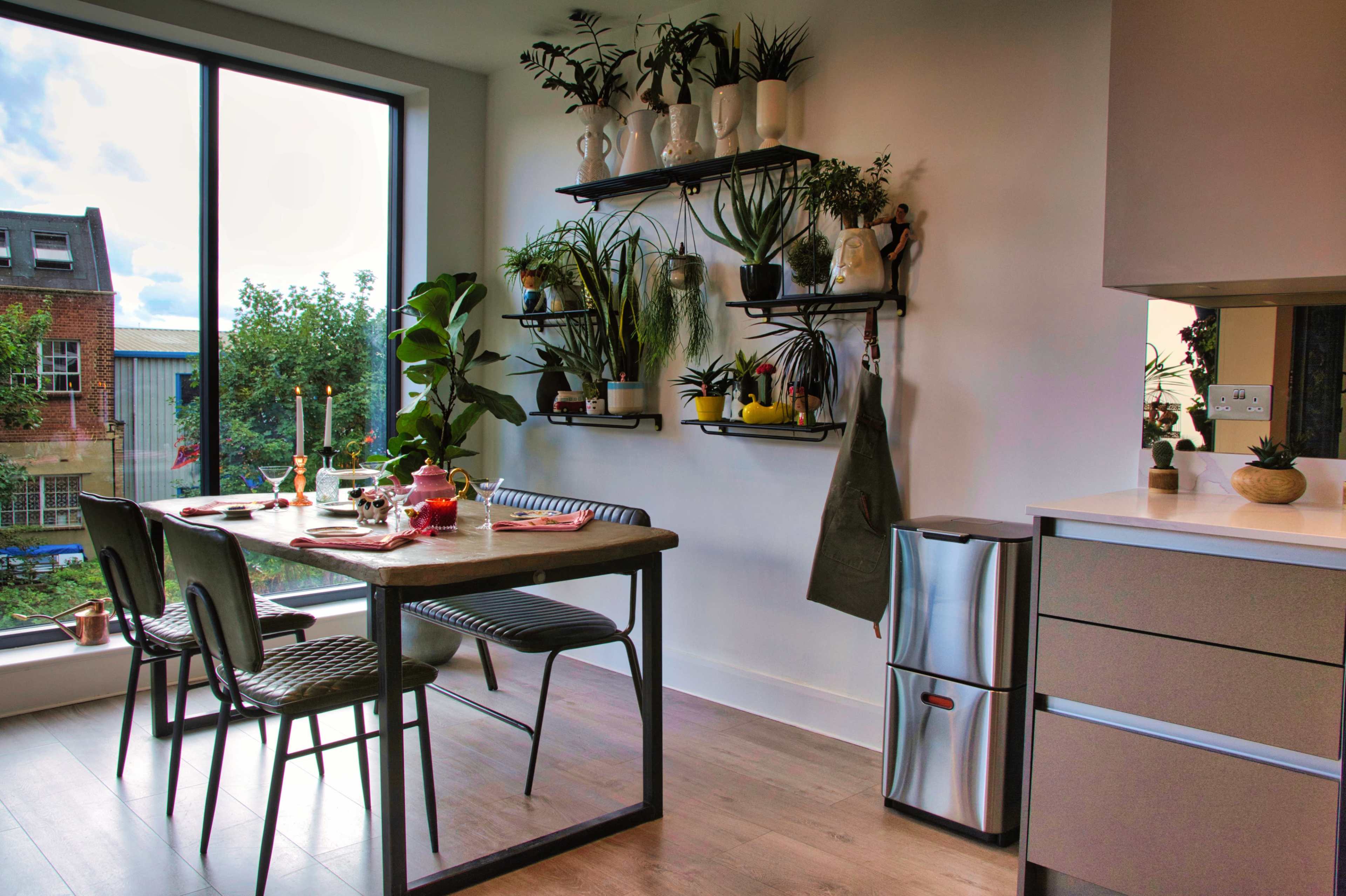 A modern kitchen features a wooden dining table surrounded by black chairs and a wall shelf with various plants and decorative items.