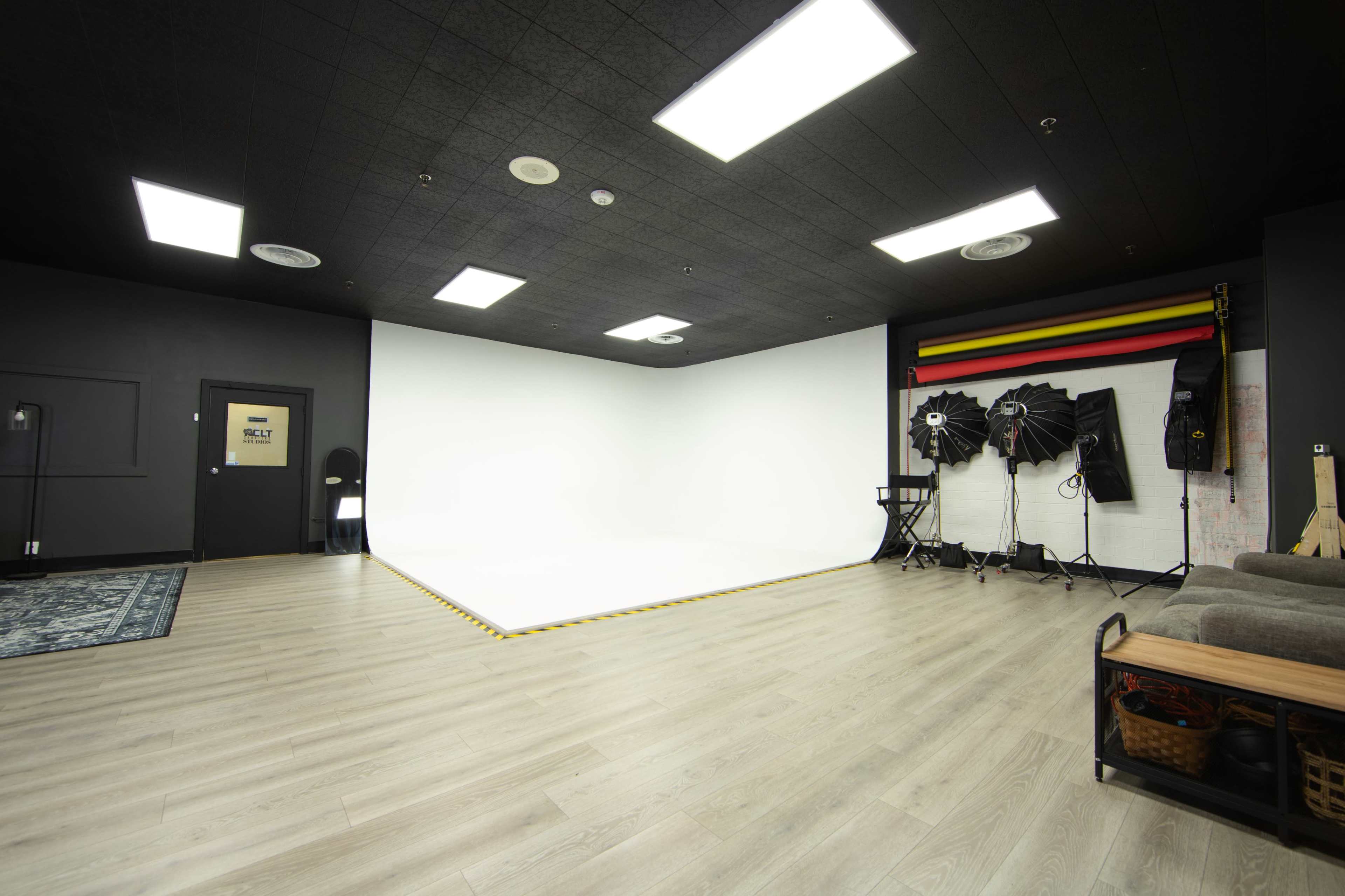 The image shows a spacious photography studio with a large white backdrop, several lighting equipment setups, and wooden flooring.