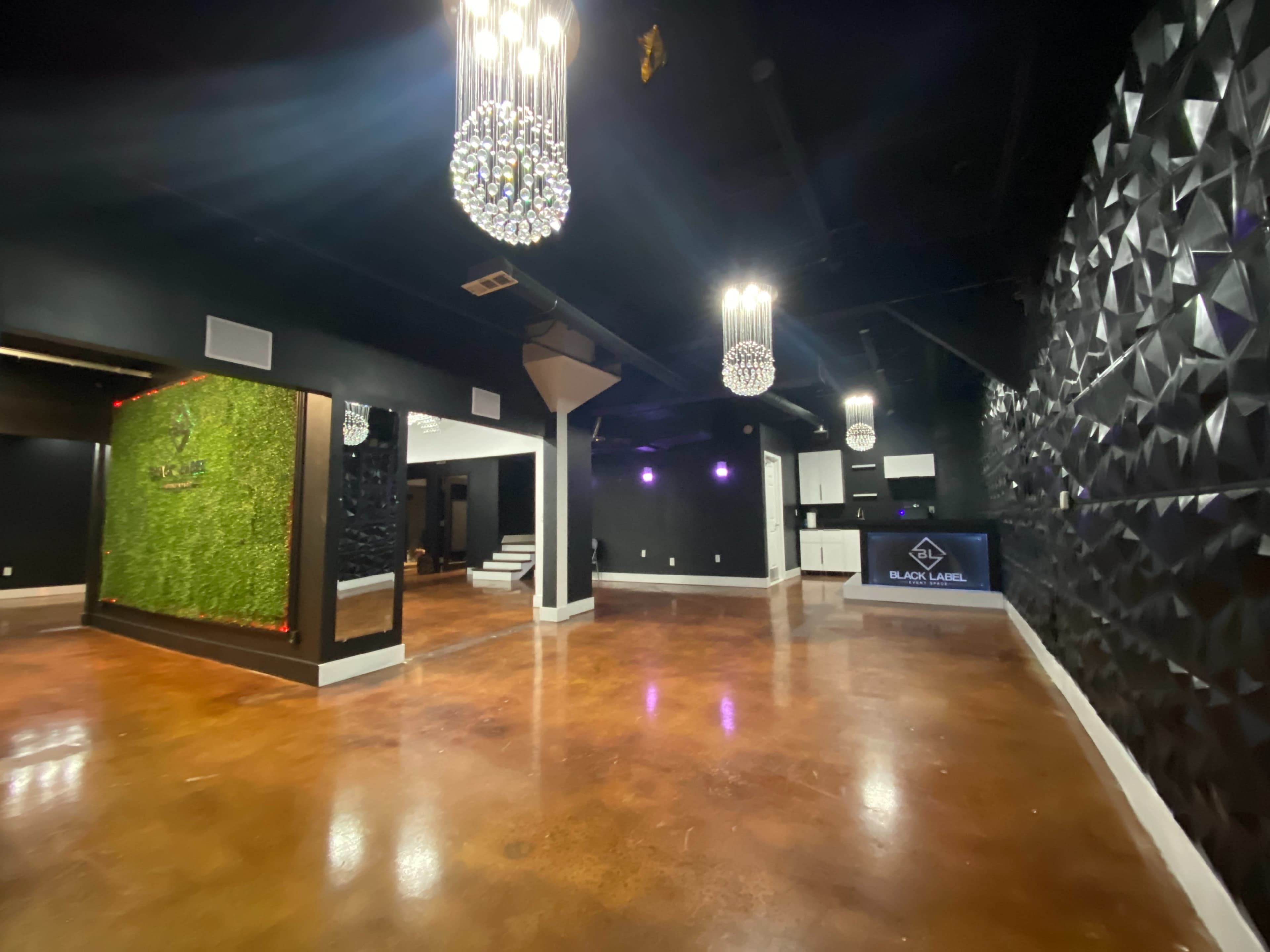 The image shows an empty, contemporary event space featuring black walls, decorative greenery, and pendant chandeliers.
