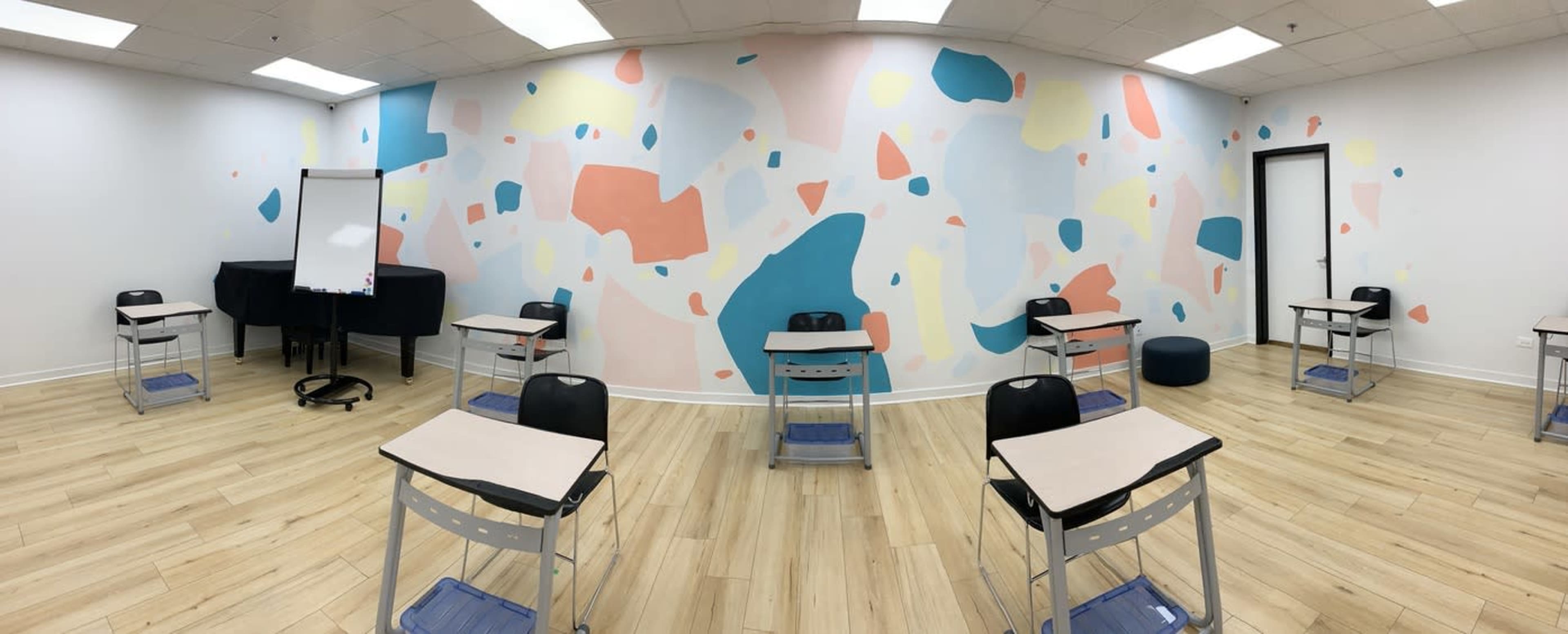 The image shows a modern classroom with several black chairs and desks arranged in a circular layout, featuring a colorful abstract mural on the walls.