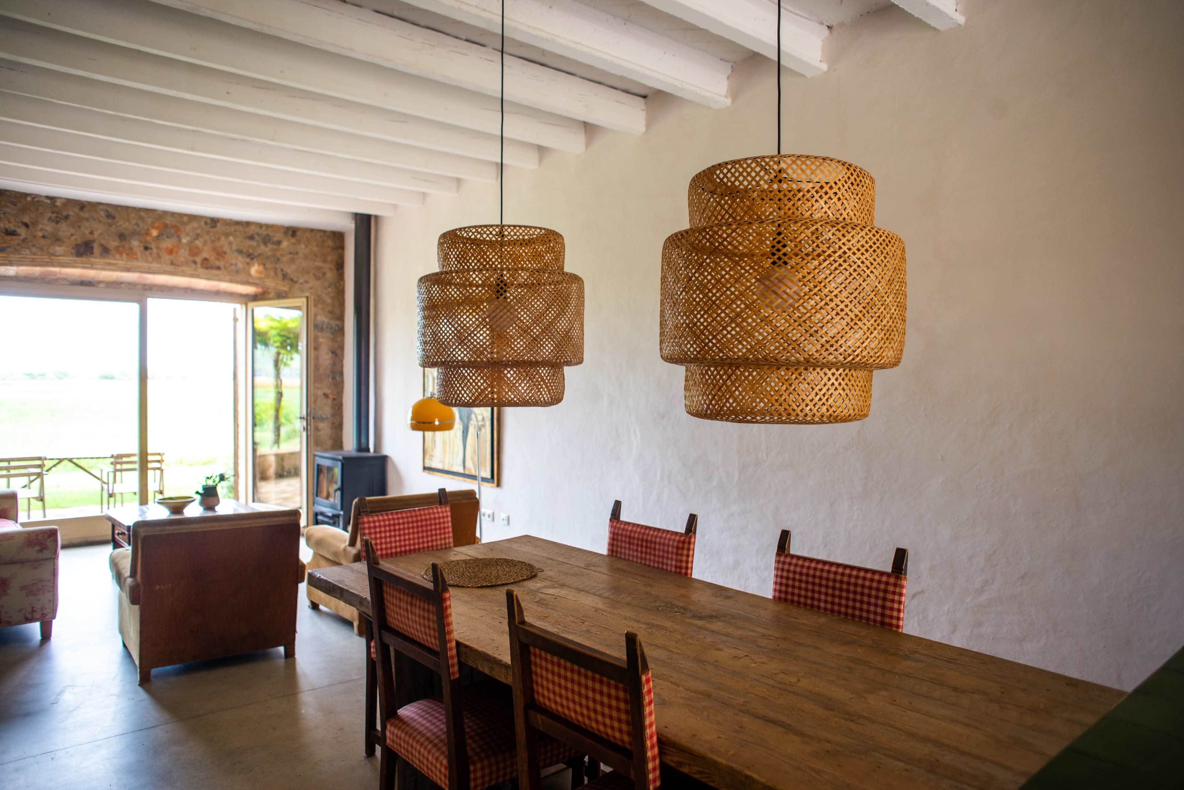 A dining area features a wooden table surrounded by chairs, with woven pendant lights hanging above and large windows showing a view of the outdoors.