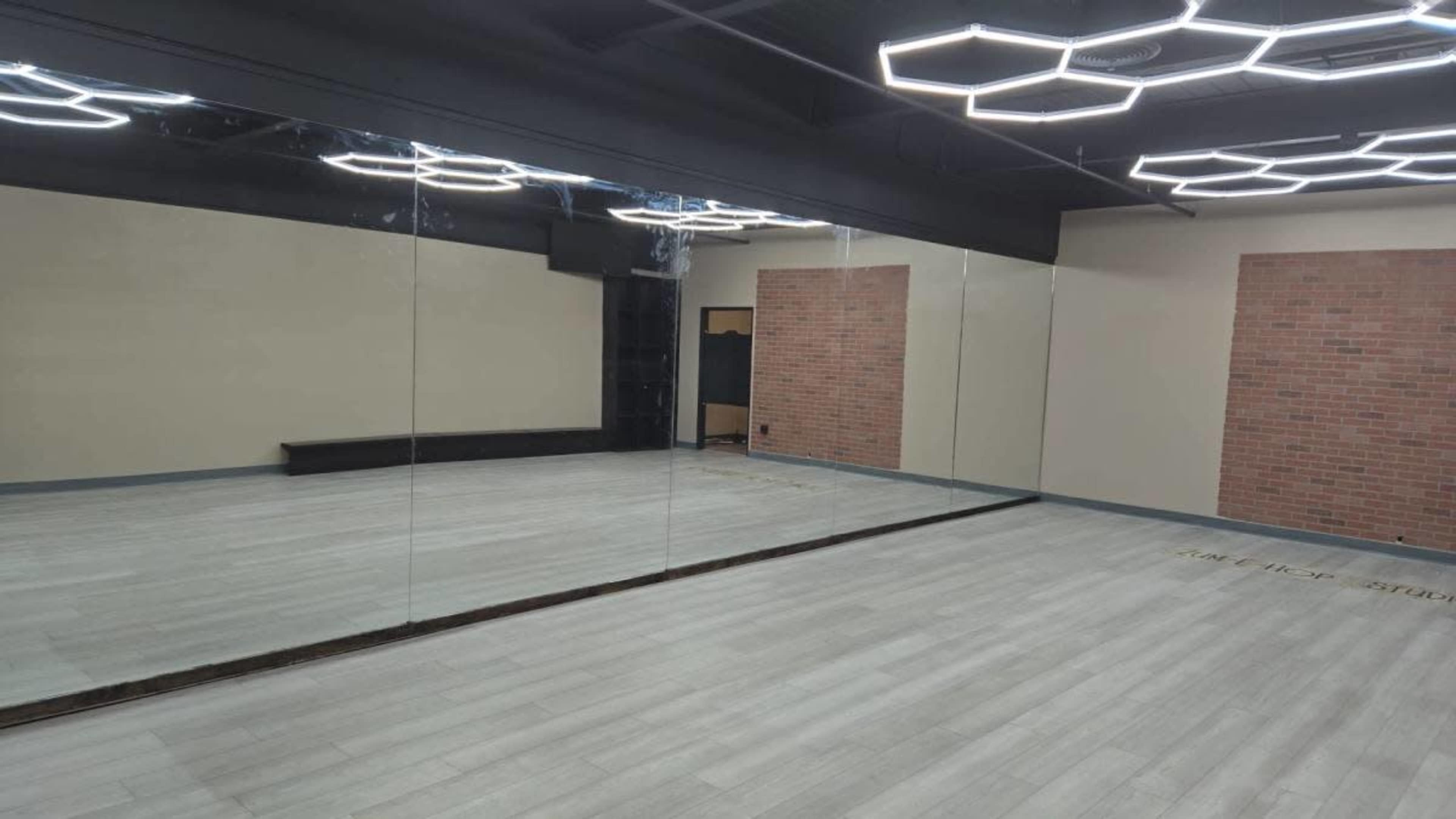 The image shows a spacious, empty studio room with mirrored walls, wooden flooring, and modern lighting fixtures on the ceiling.