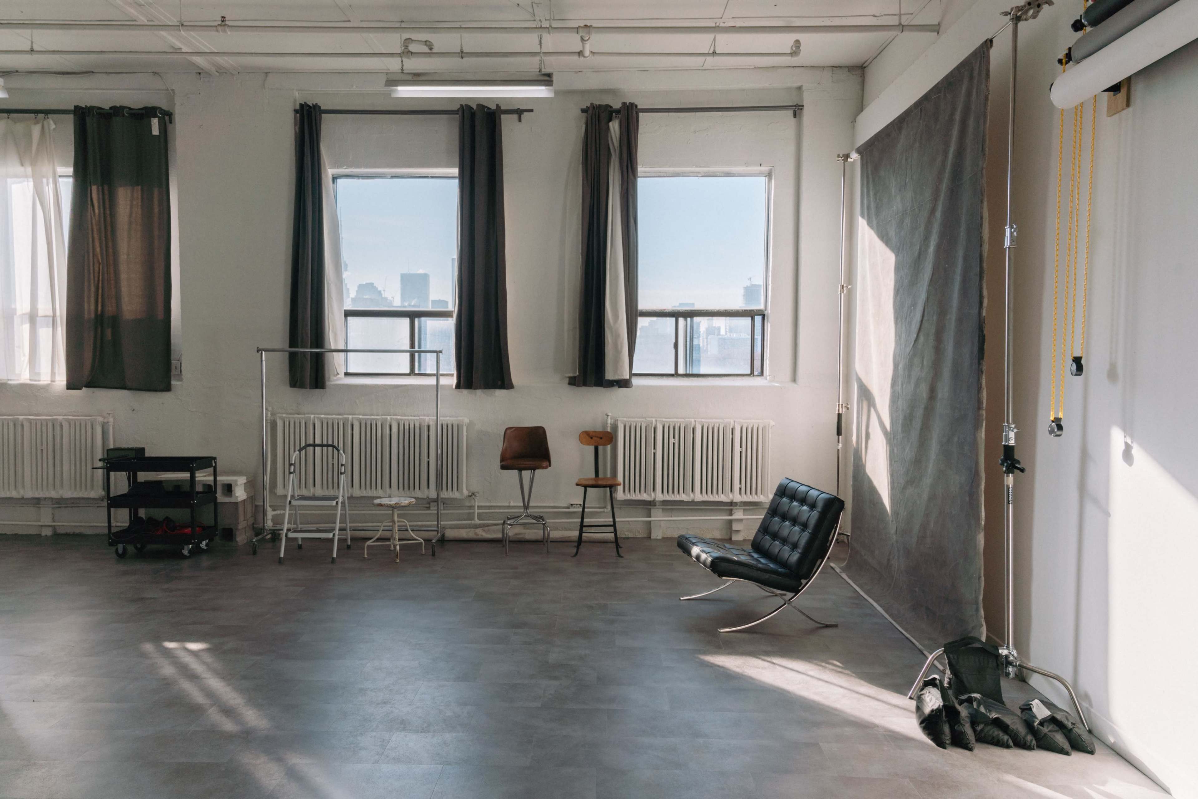 A spacious, sunlit studio features large windows, a minimalist black chair, and various pieces of furniture along the walls.