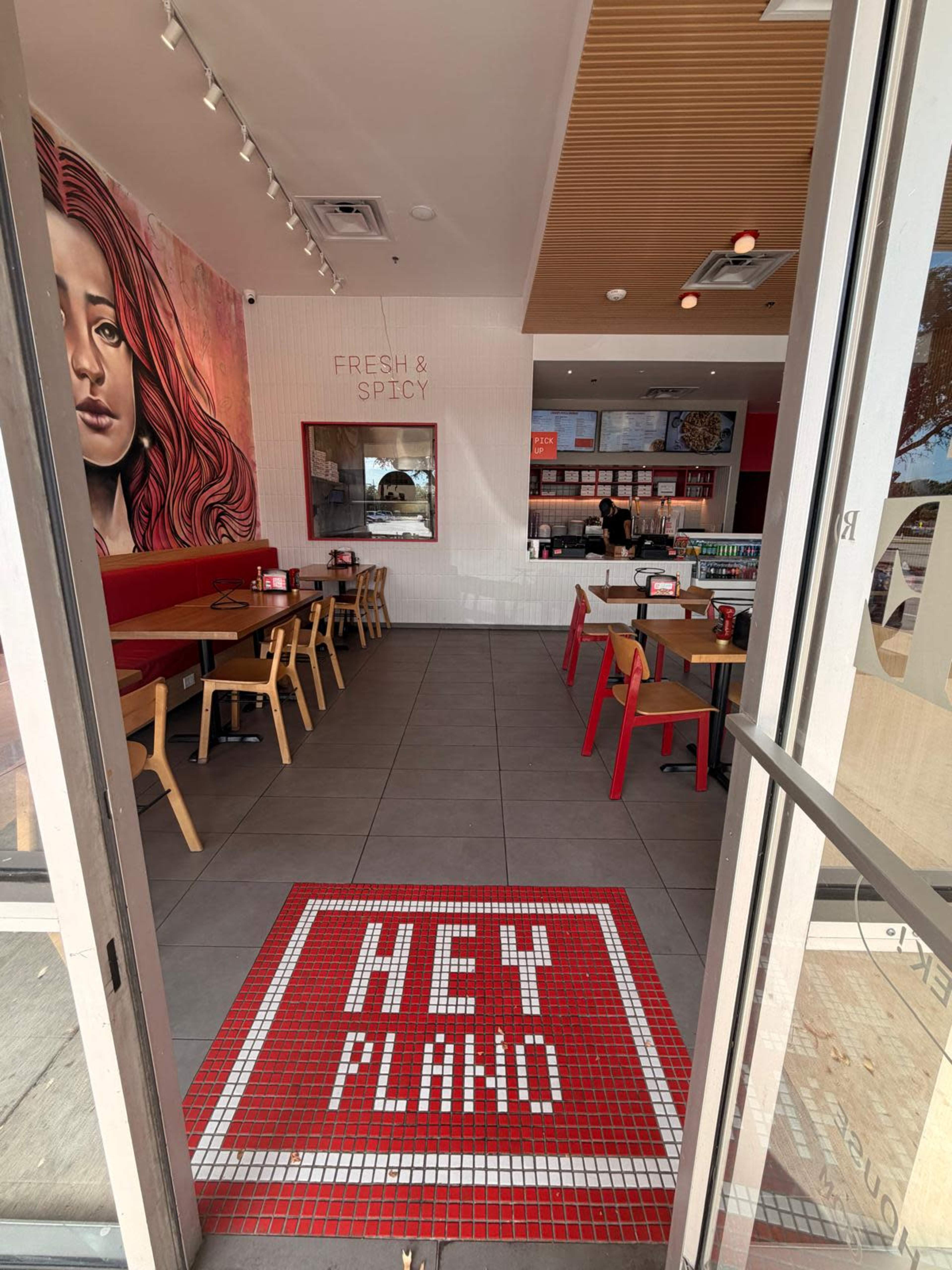 The image shows the interior of a restaurant with red and wooden furniture, featuring a large mural on one wall and a welcome mat that reads "HEY PLANO" at the entrance.
