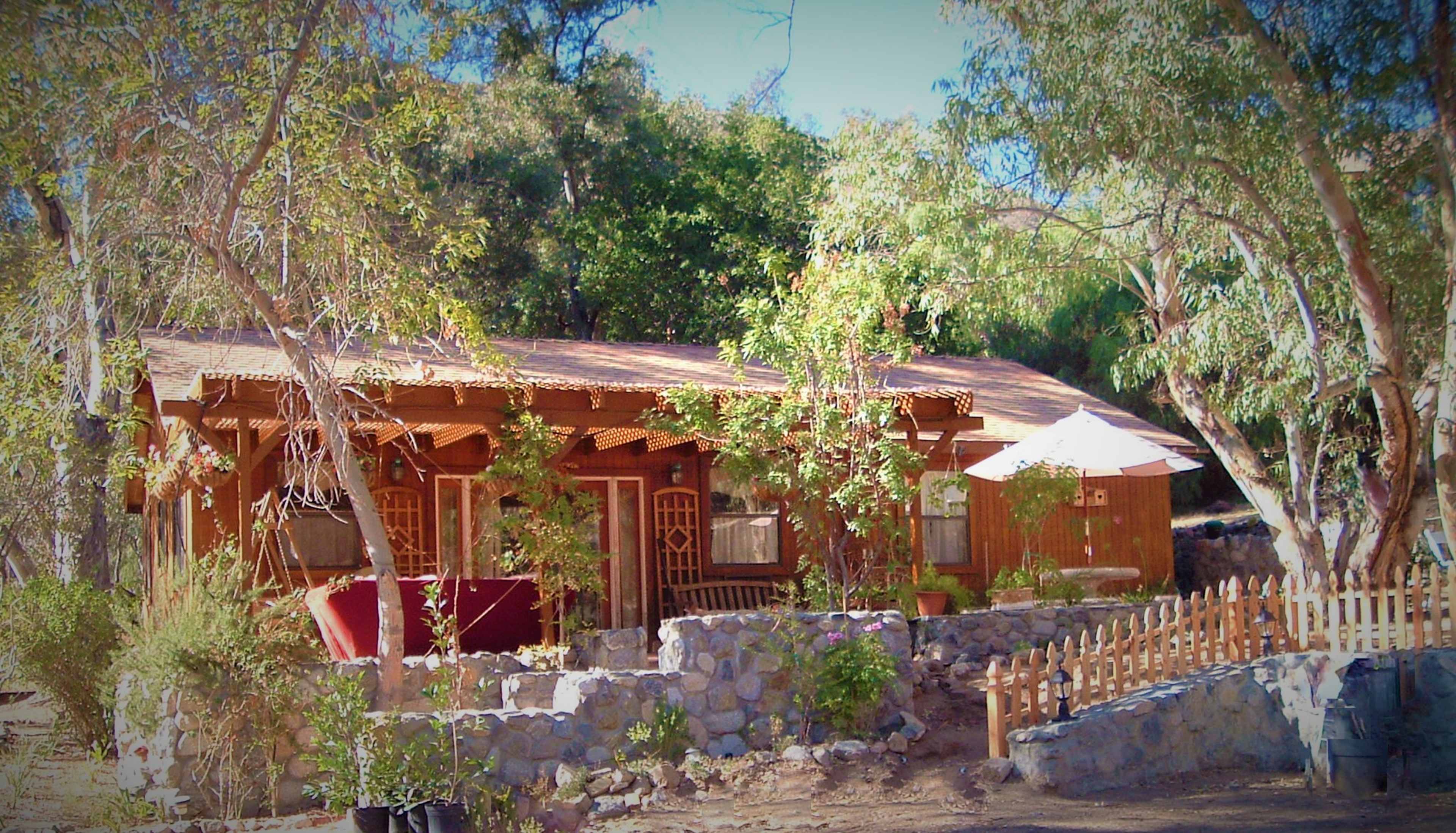 A wooden cabin with a stone wall and wooden fence is surrounded by trees and shrubs.