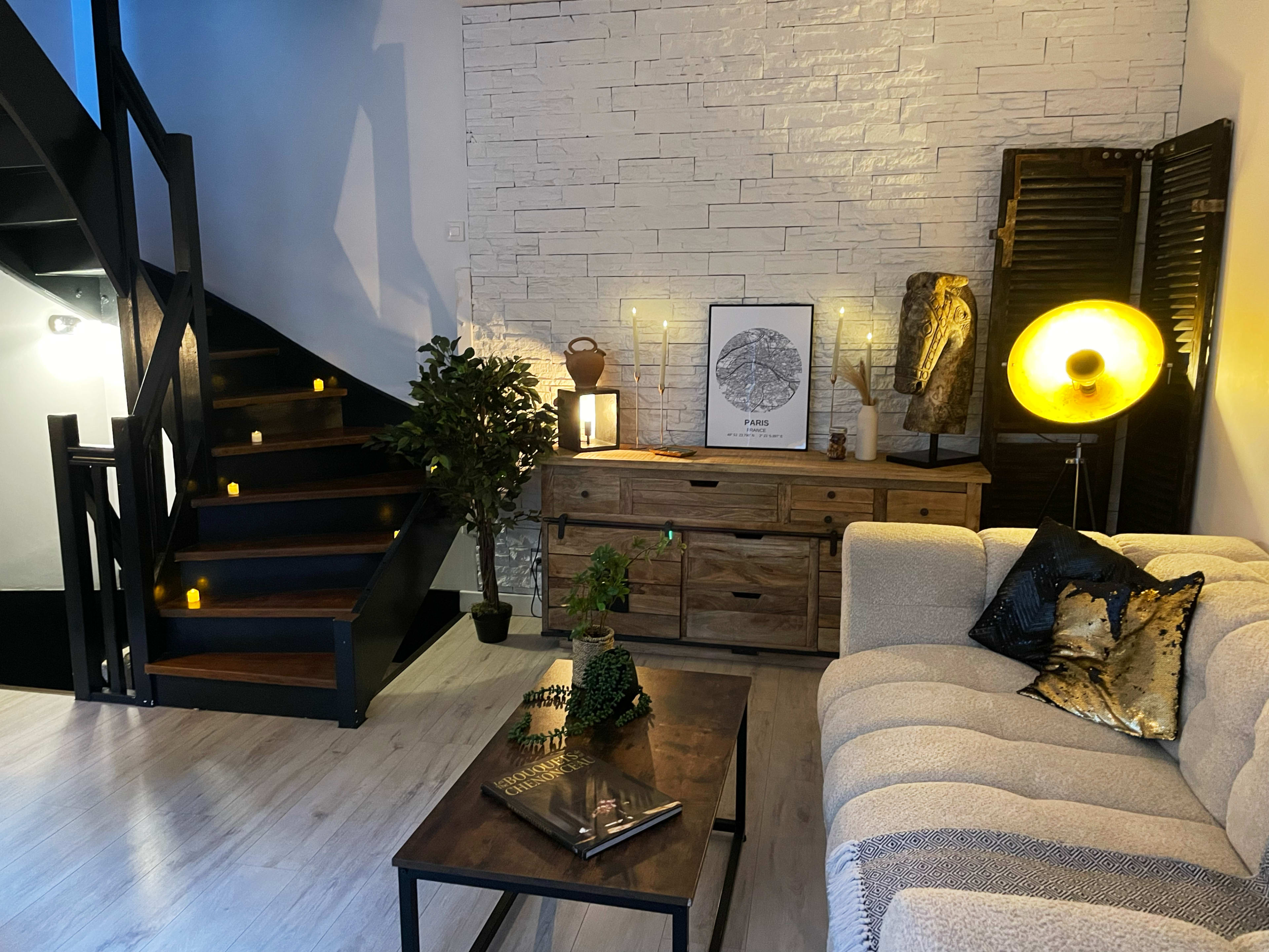 The image shows a modern living room with a staircase, a wooden chest of drawers, a couch, and decorative elements including plants and lighting.