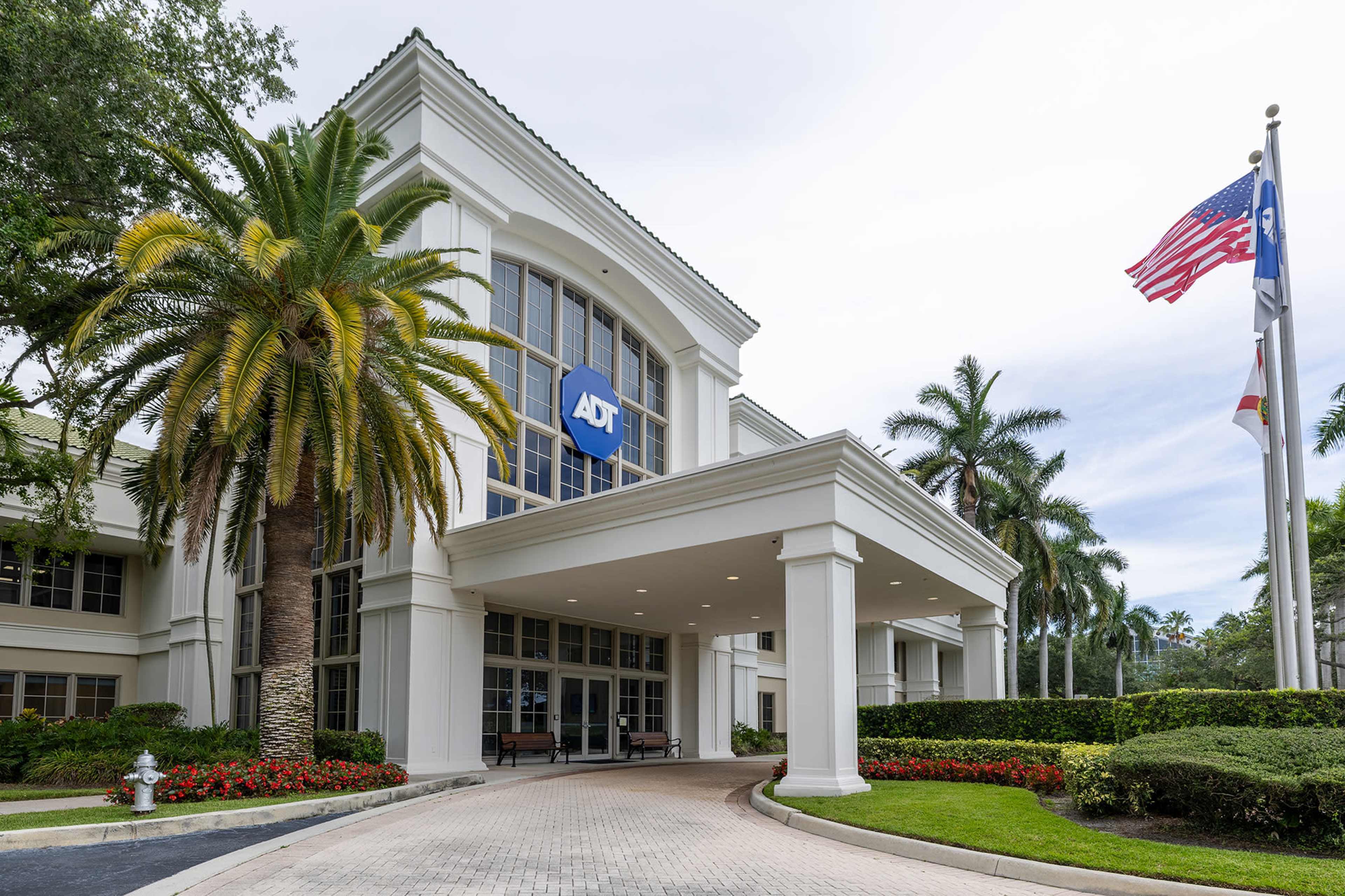 The image shows the entrance of the ADT building, featuring a large logo, palm trees, and flags outside.