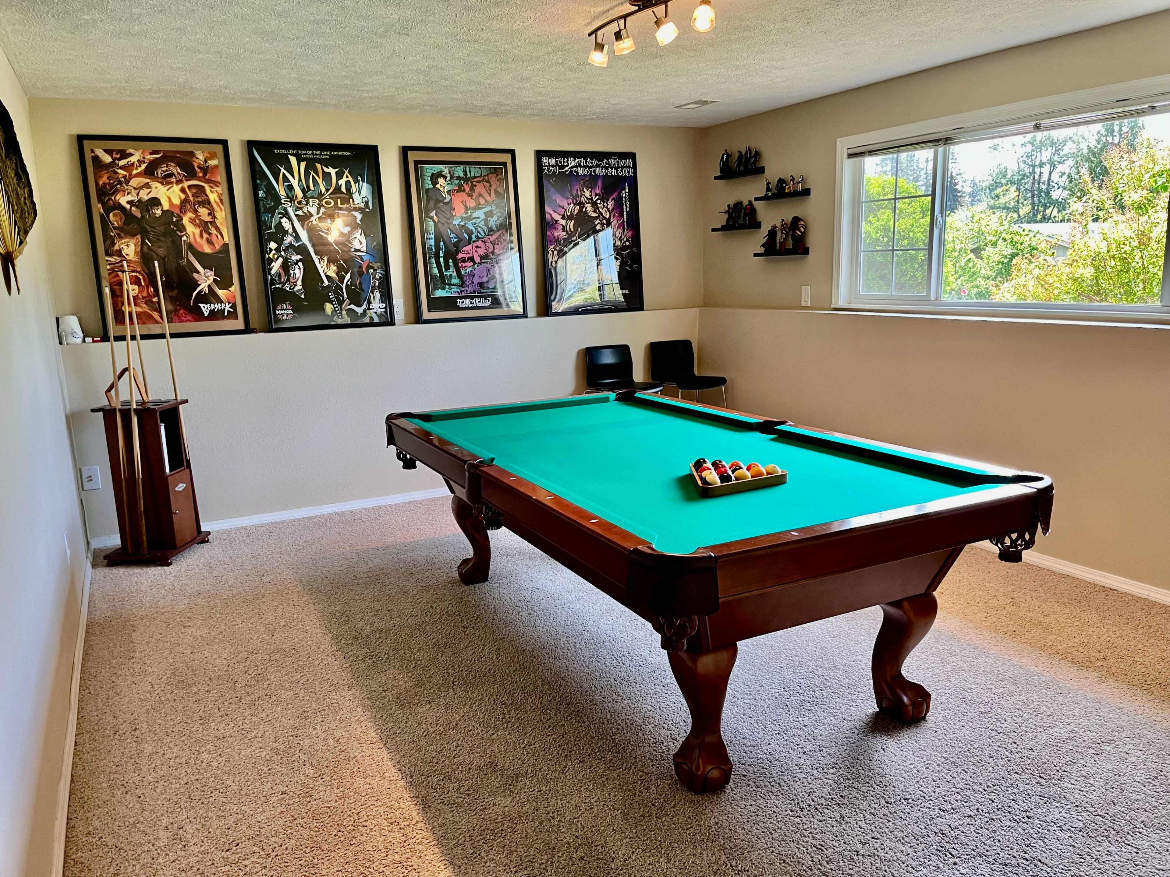 A game room with a green felt pool table, several framed posters on the wall, and a window overlooking greenery outside.
