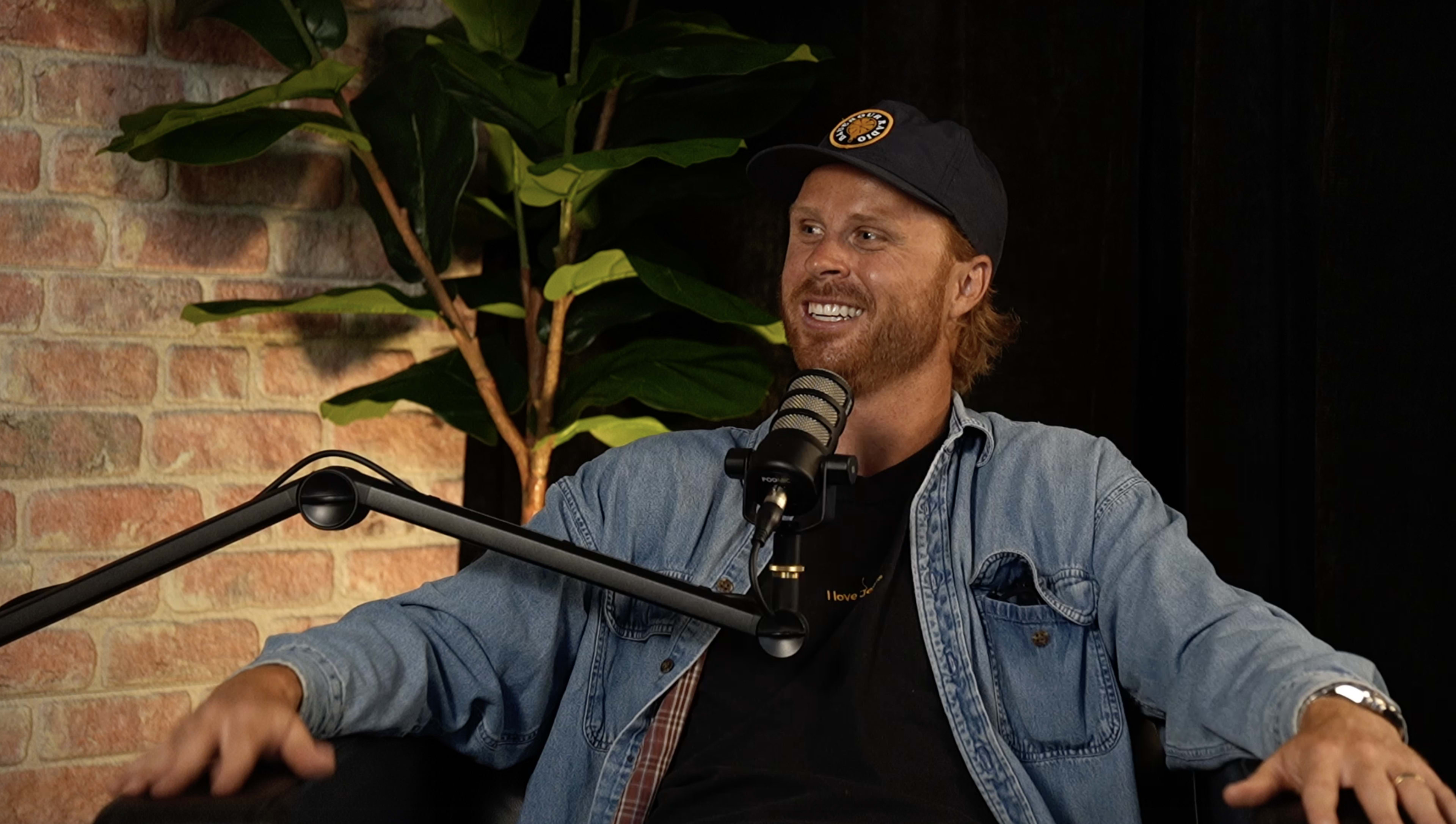 A man in a denim jacket and baseball cap sits comfortably in a studio setting with a microphone in front of him and a leafy plant in the background.