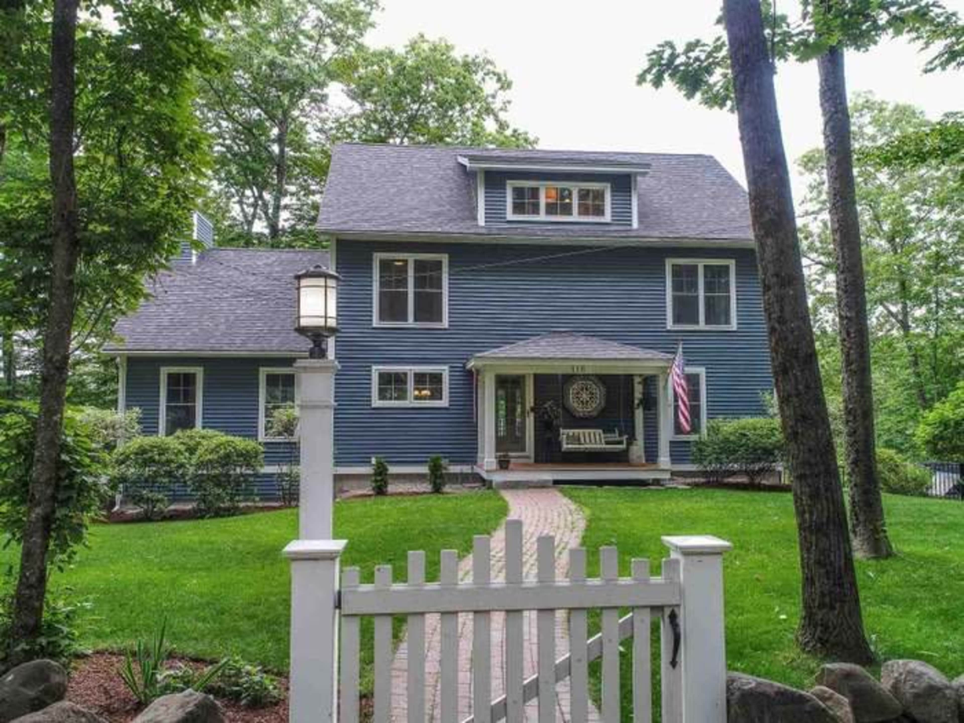 A blue two-story house with a front porch and a white picket fence is surrounded by trees and a manicured lawn.