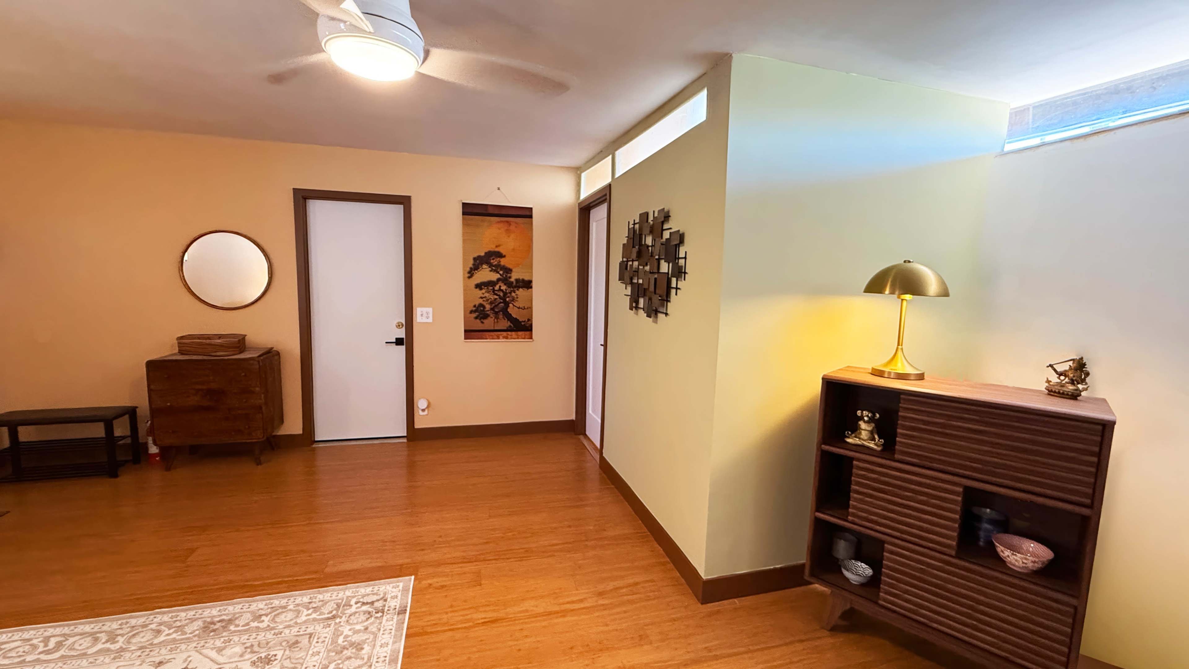 The image shows a warmly lit, minimalist room featuring a wooden floor, a small console table, a round wall mirror, and a decorative wall hanging.