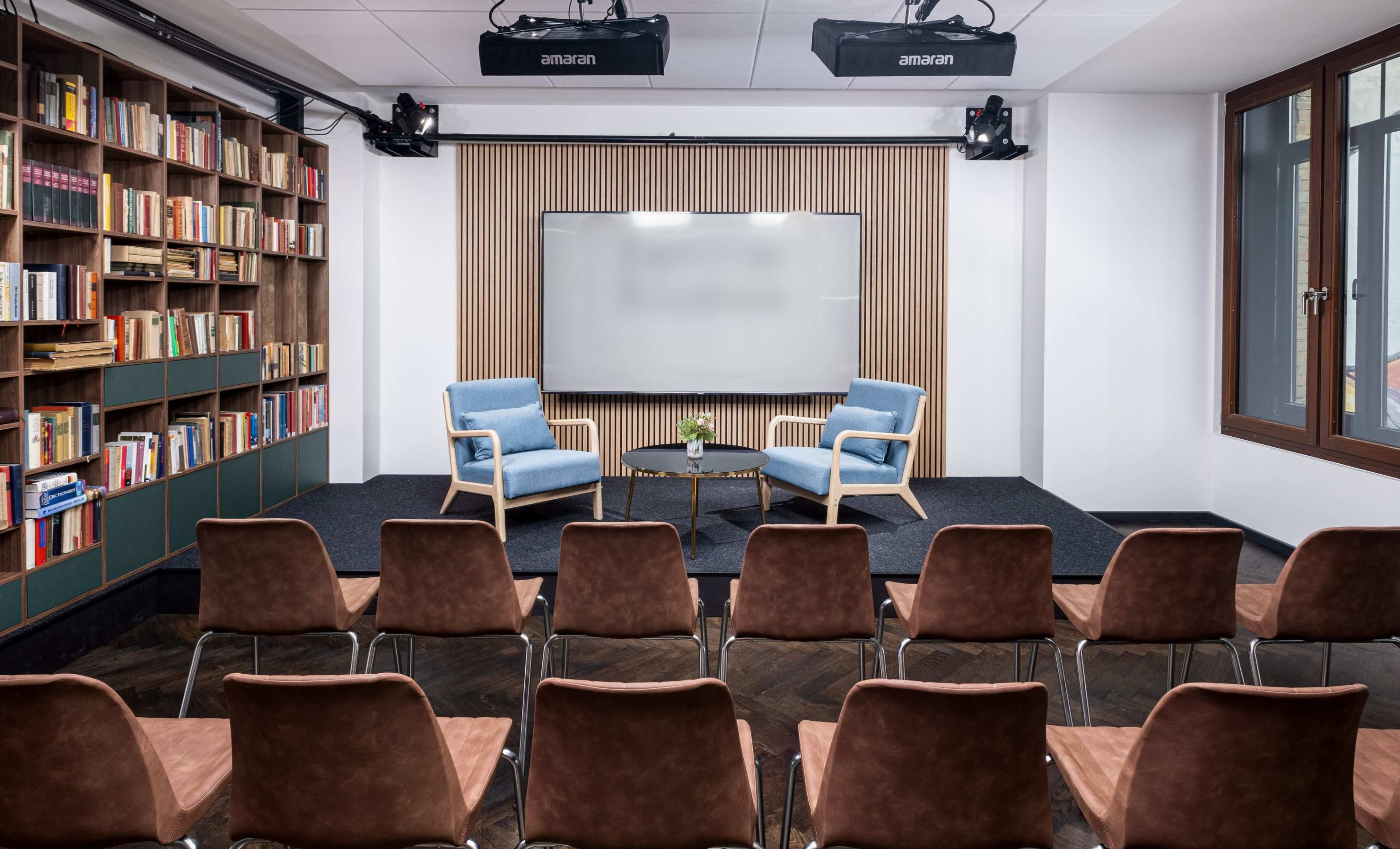 The image shows a modern seminar room with two blue chairs facing a central table, surrounded by rows of brown chairs and a wooden bookshelf filled with books.