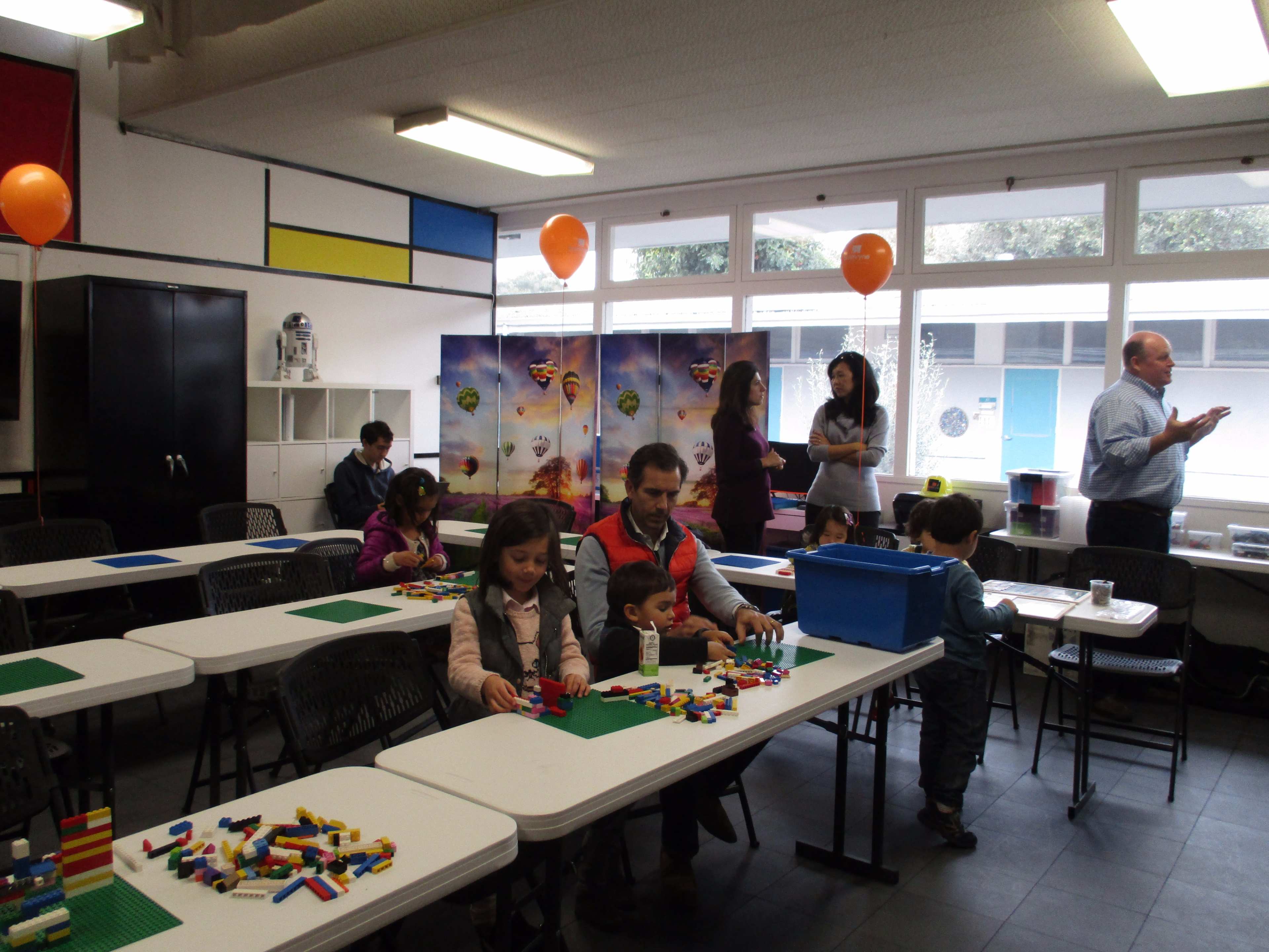 A group of children and adults are working with building blocks at tables in a classroom setting with large windows and balloons.
