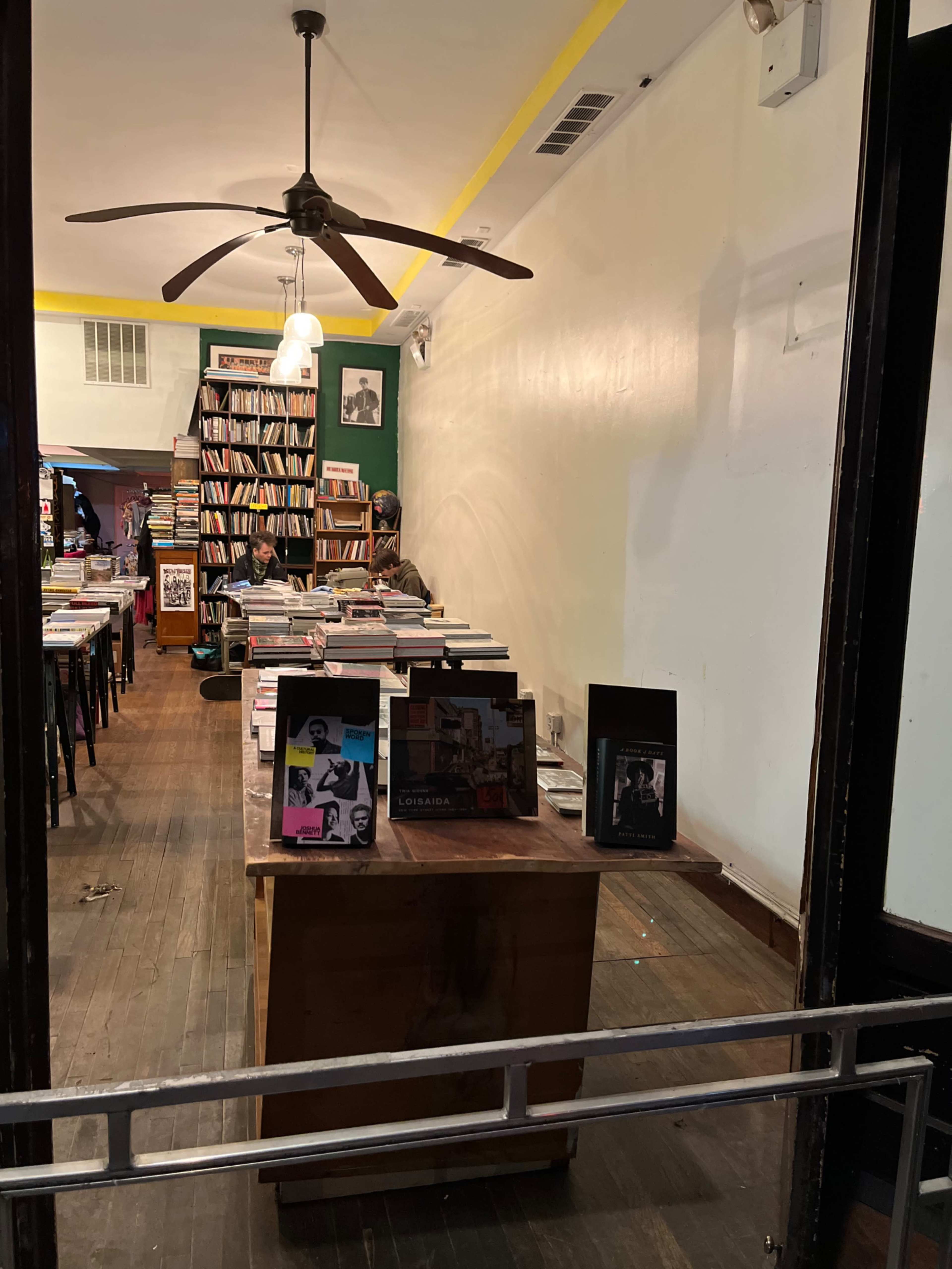 The interior of a bookstore with wooden floors, a long table displaying books, and a bookshelf filled with various titles in the background.