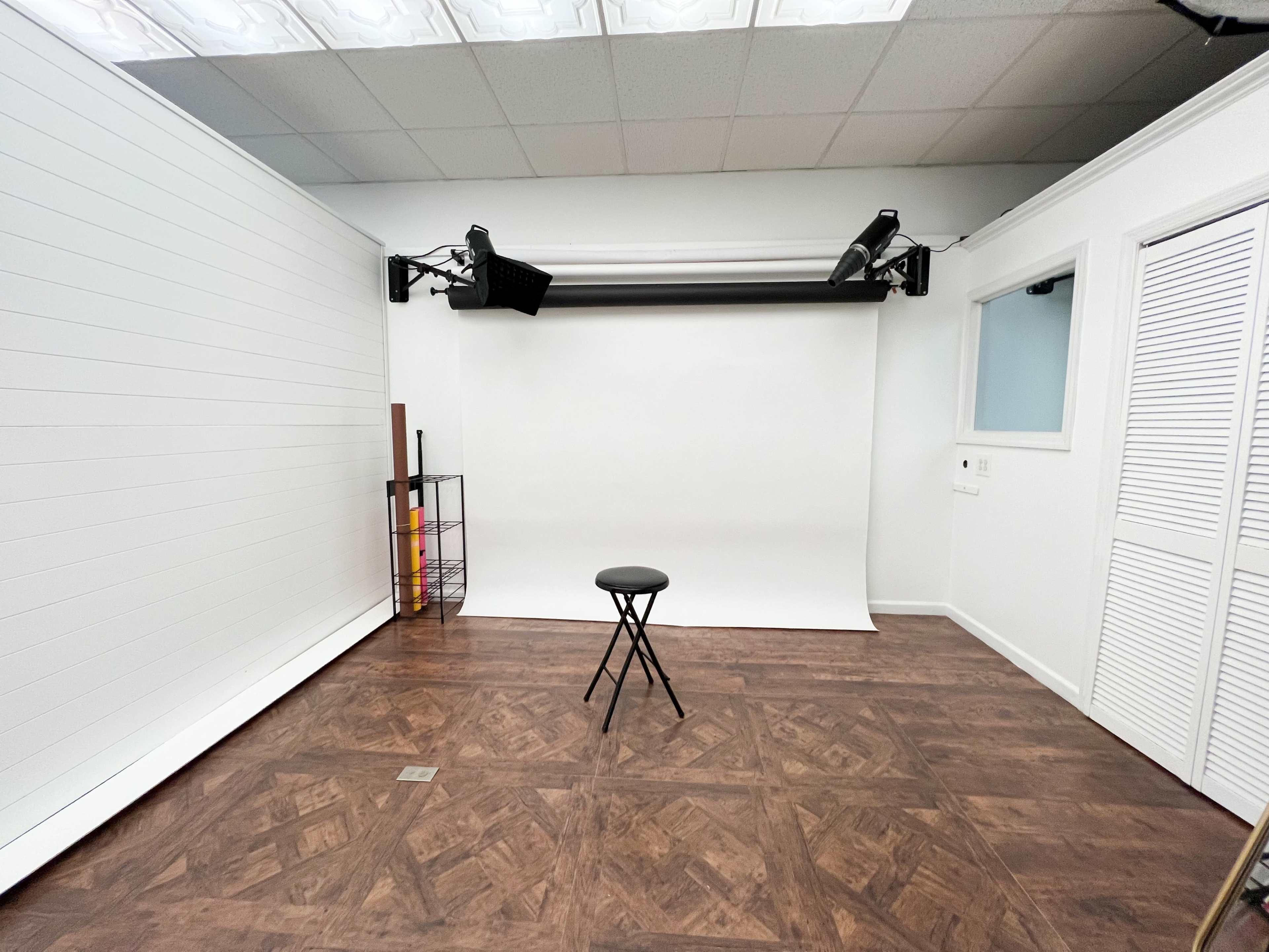 A photography studio featuring a simple setup with a plain white backdrop, a black stool in the center, and minimal equipment mounted on the ceiling.
