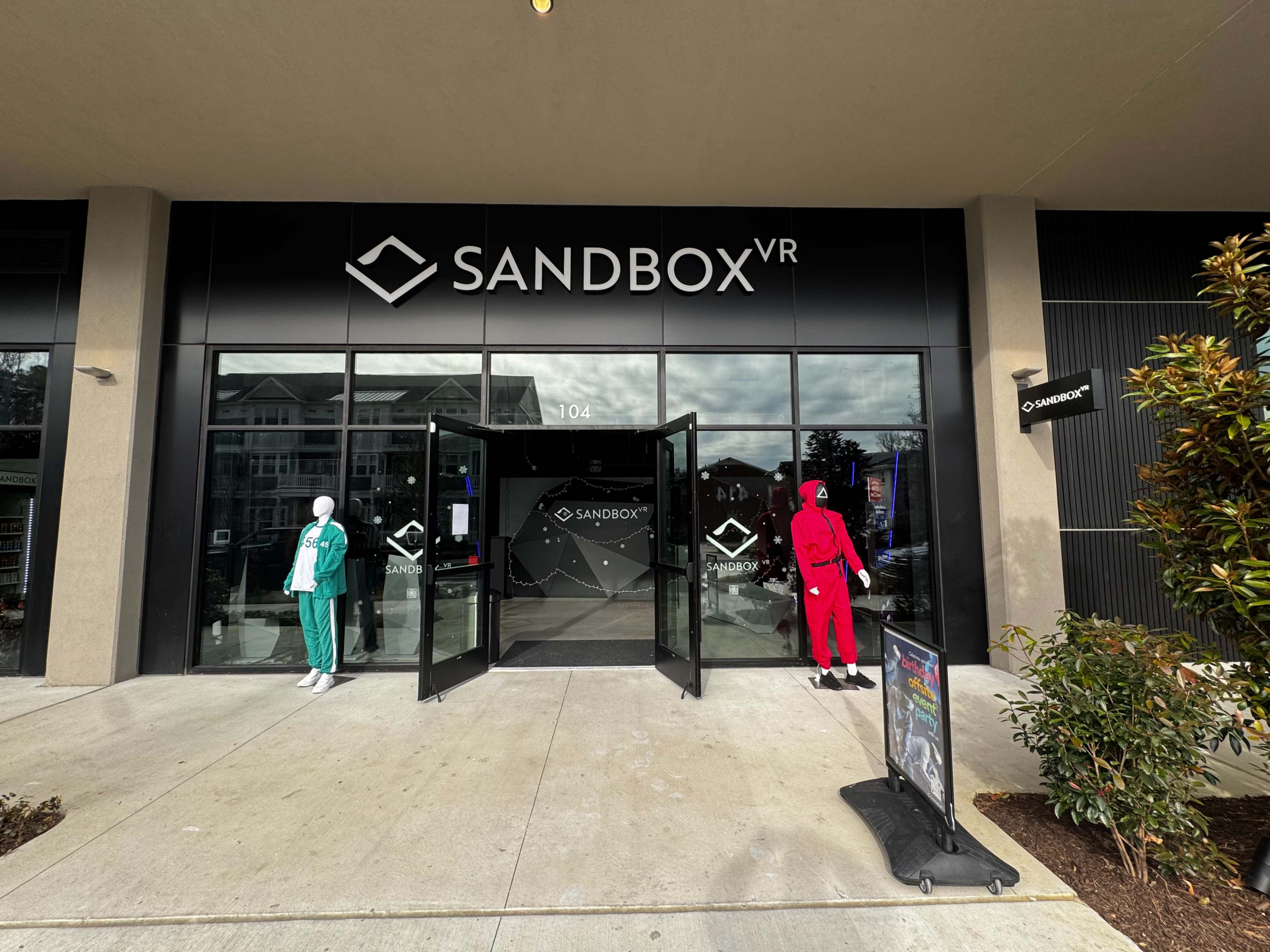 The image shows the entrance of a business called "SANDBOX VR," with two mannequins dressed in colorful outfits standing outside the door.