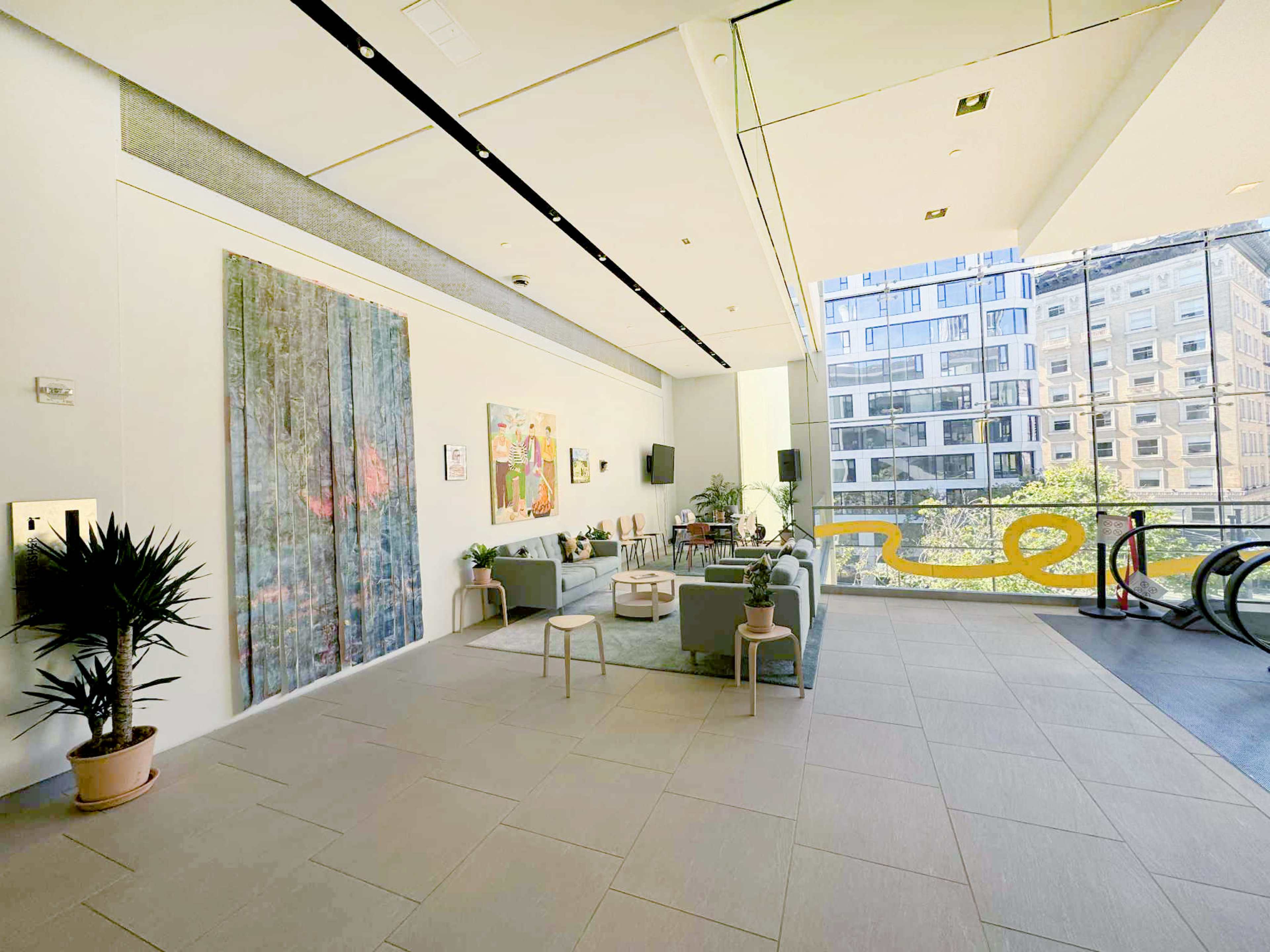The image shows a modern, open lobby area featuring artwork on the wall, seating arrangements, and large windows looking out onto a cityscape.