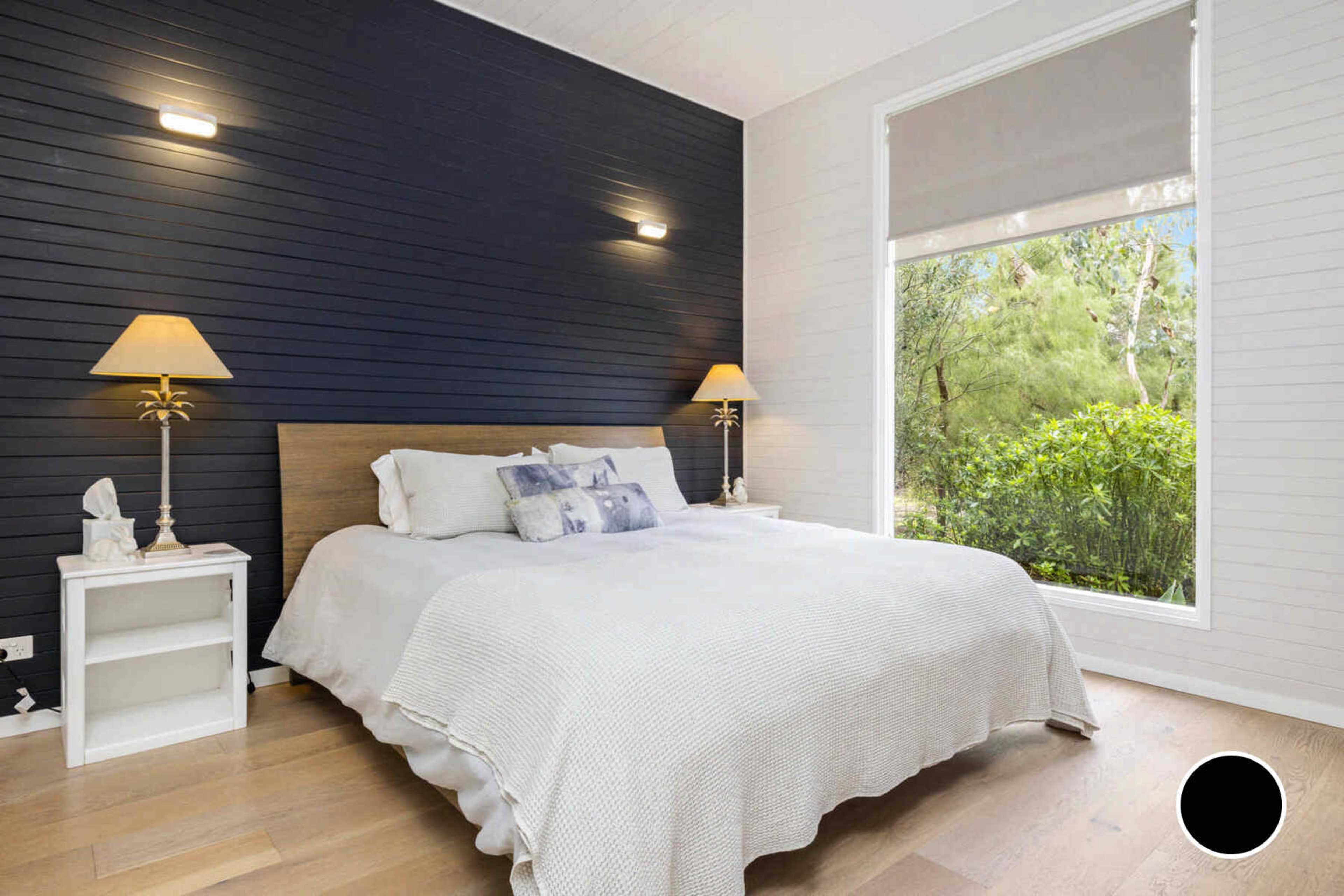 The bedroom features a bed with a light-colored blanket against a dark accent wall, two bedside lamps, and a large window offering a view of greenery.