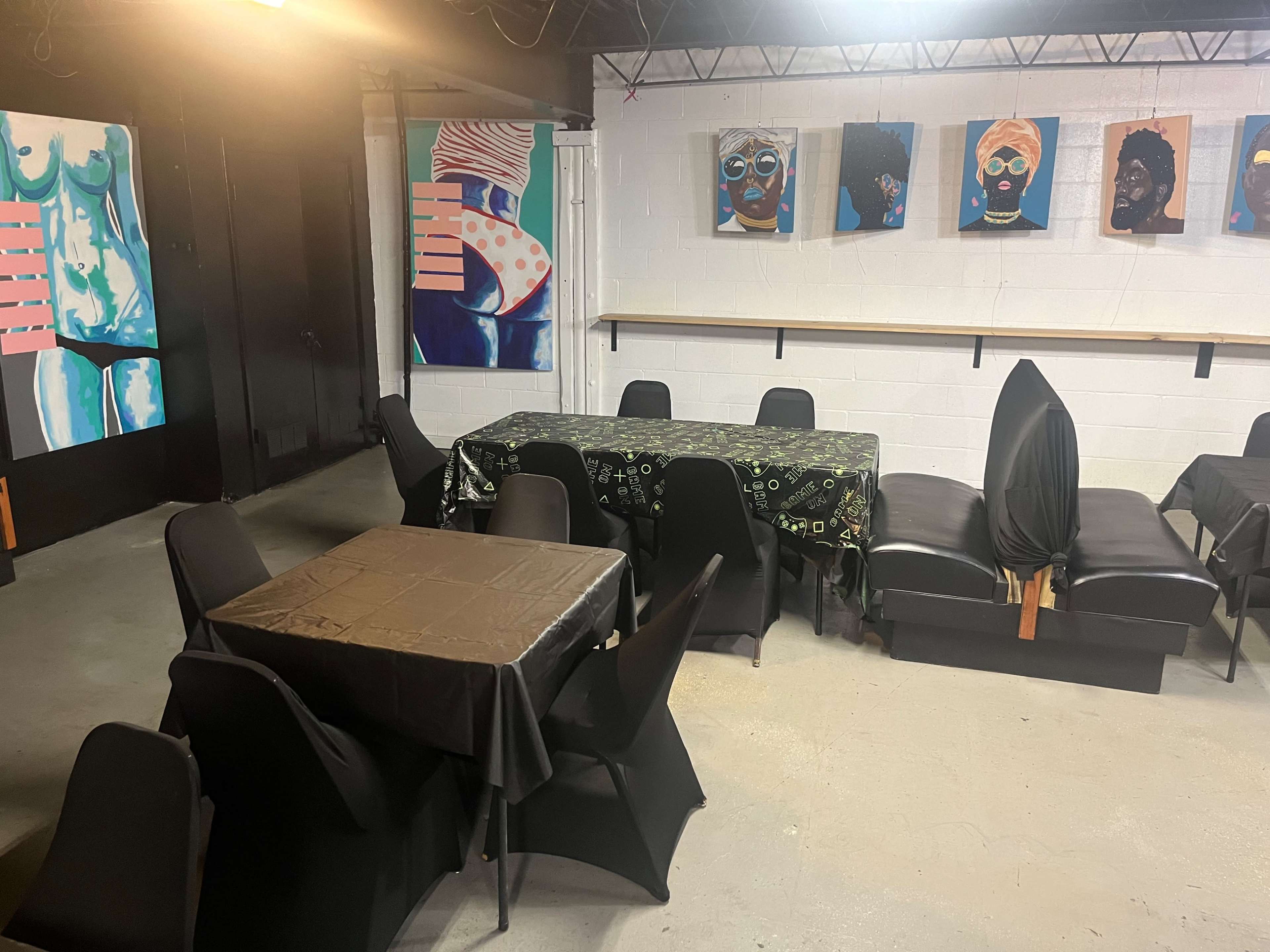 The image shows a dimly lit interior space with several tables covered in black tablecloths and colorful artwork hanging on the walls.