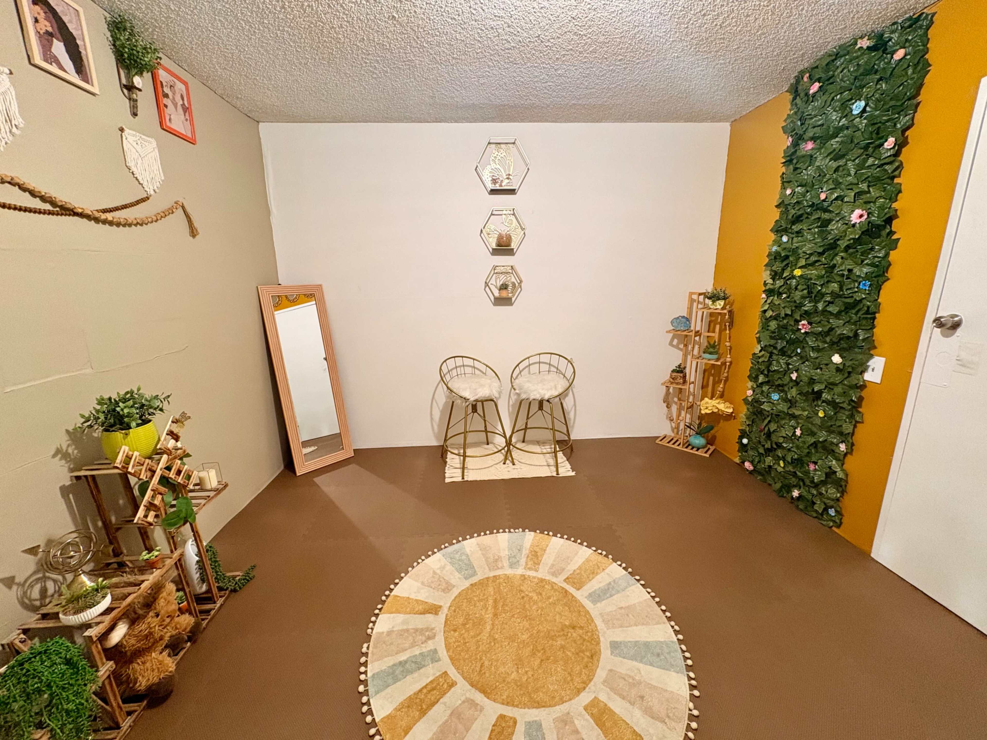 The image shows a cozy room featuring two gold chairs, a round rug with sunburst patterns, a full-length mirror, and a wall adorned with greenery and decorative shelves.