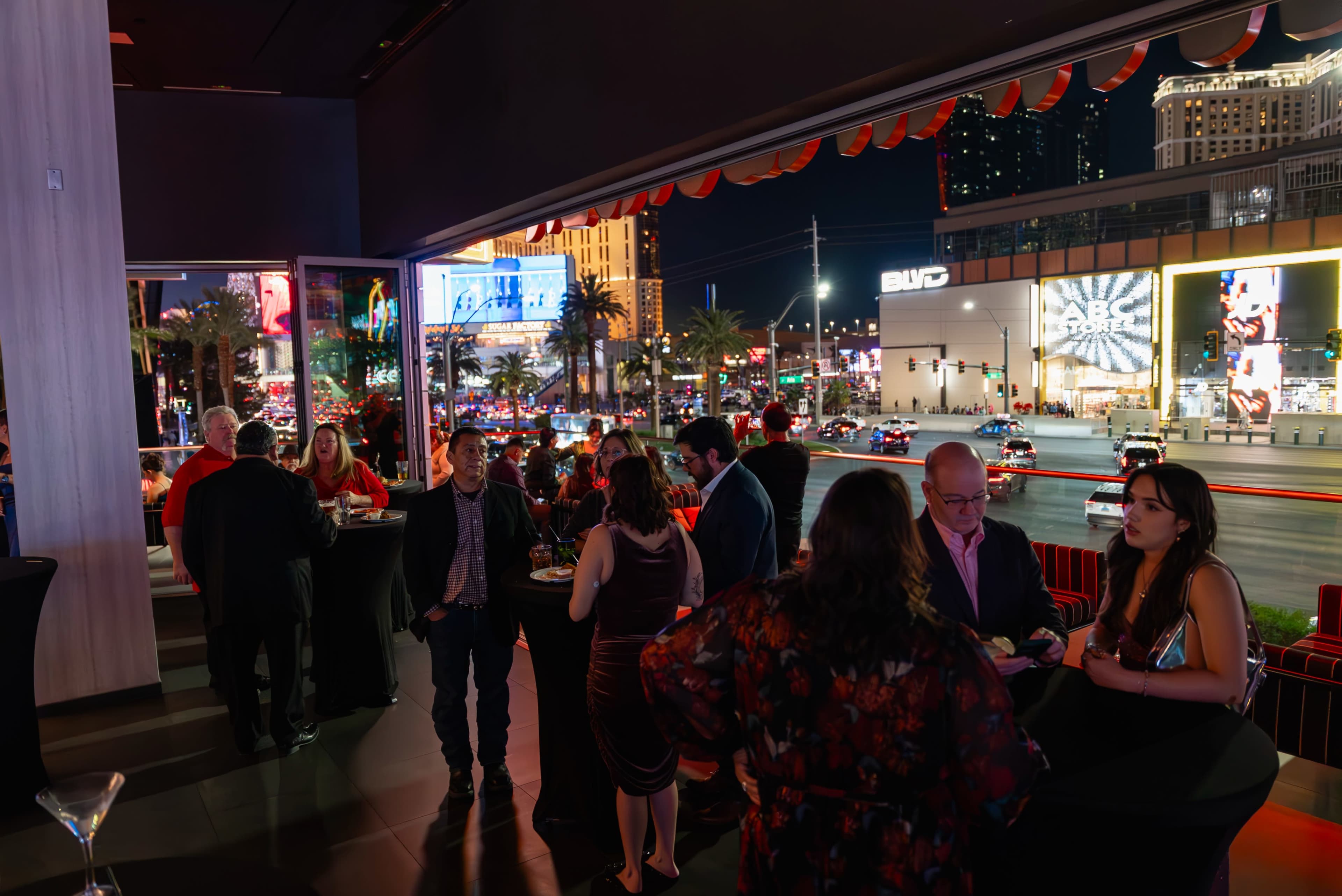 A group of people socializes at a stylish indoor event space with large windows showcasing the illuminated Las Vegas Strip at night.