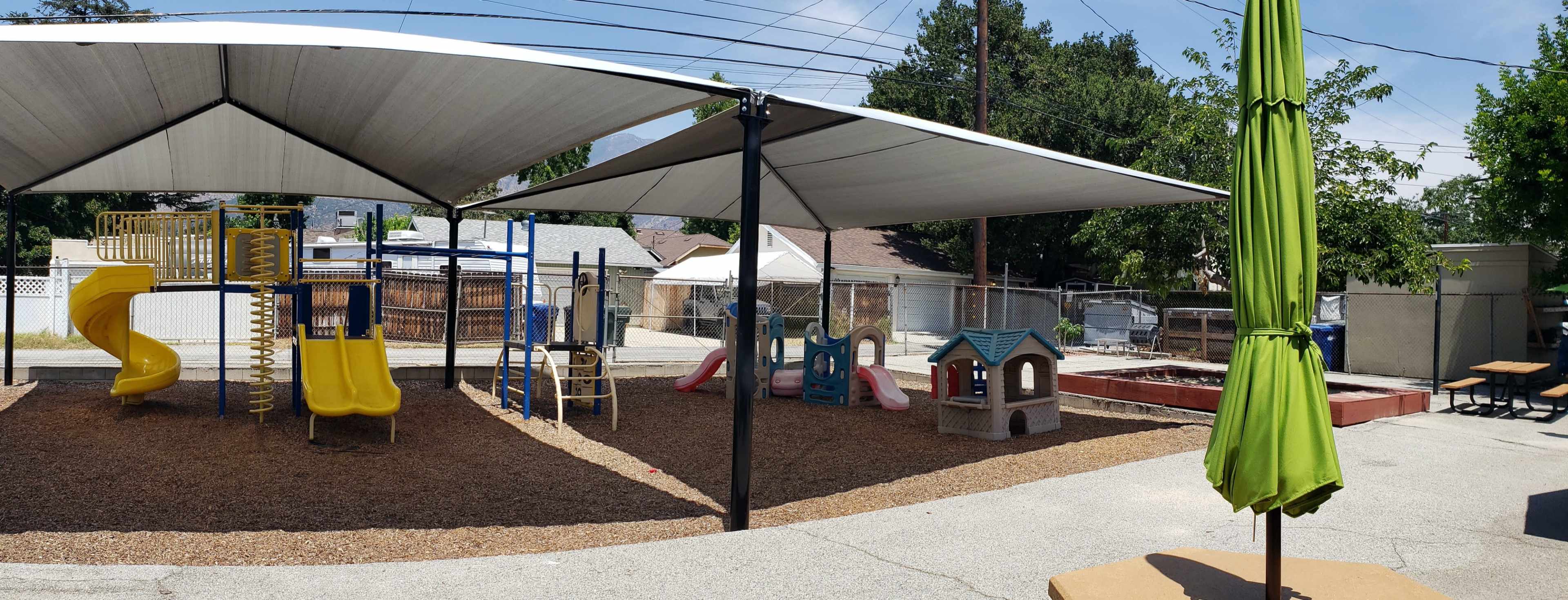The image shows a playground area with yellow slides, a small playhouse, and shaded structures surrounded by a gravel surface.