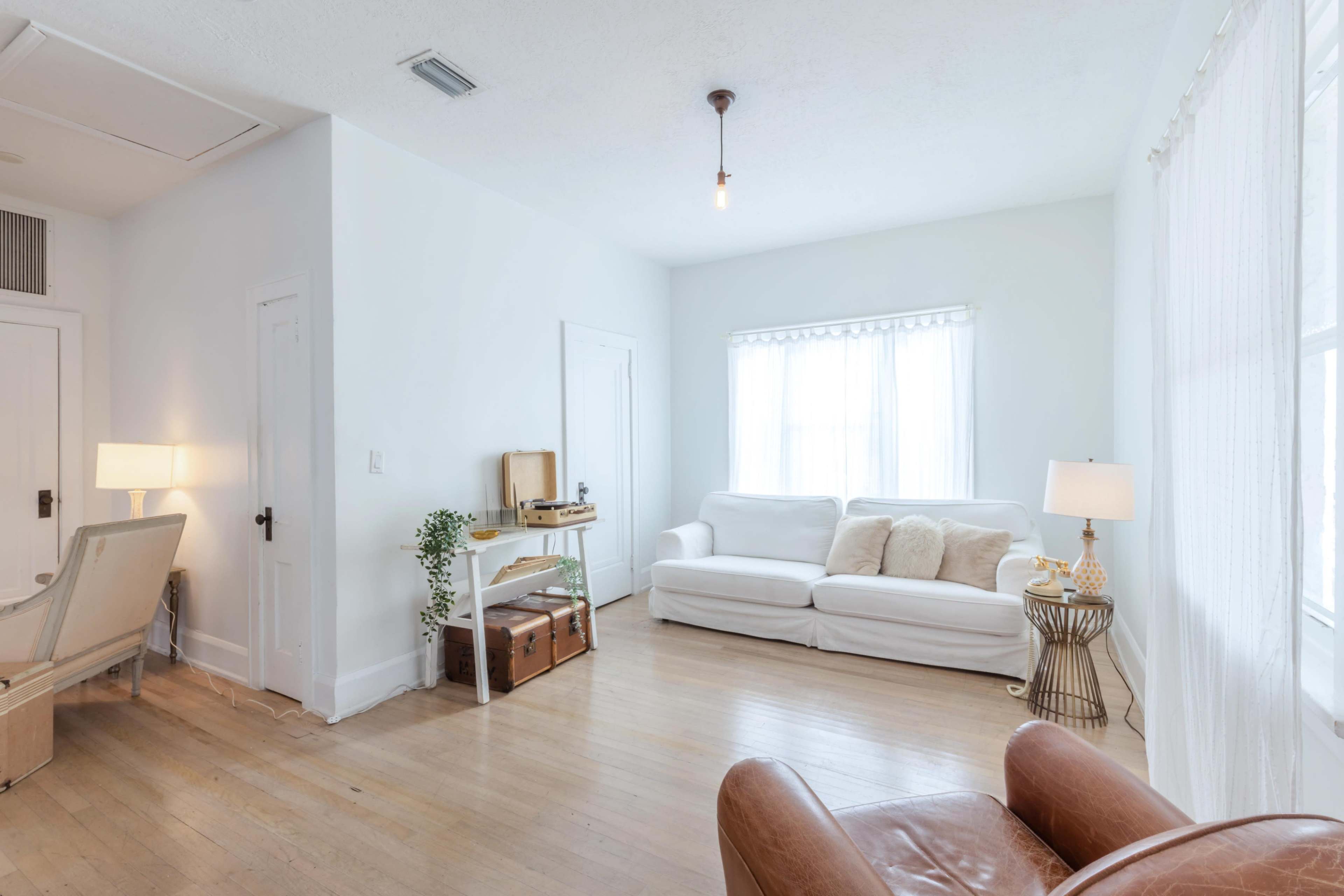 A bright living room with white walls, a white sofa, a wooden table with a plant, and a cozy chair near a window.