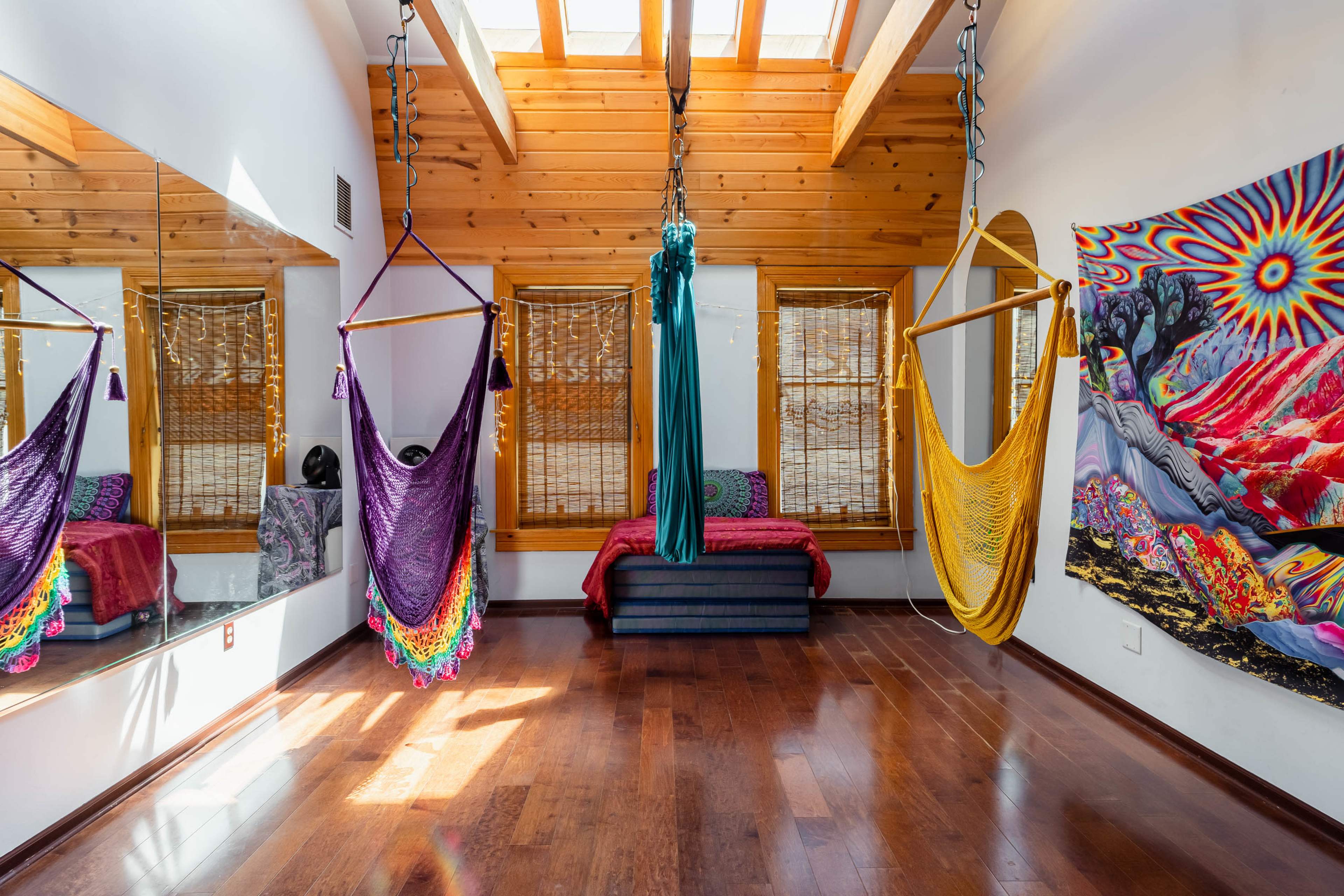 A room with wooden beams and a skylight, featuring colorful hammocks and a large tapestry on the wall, alongside a mirrored surface and rustic furnishings.