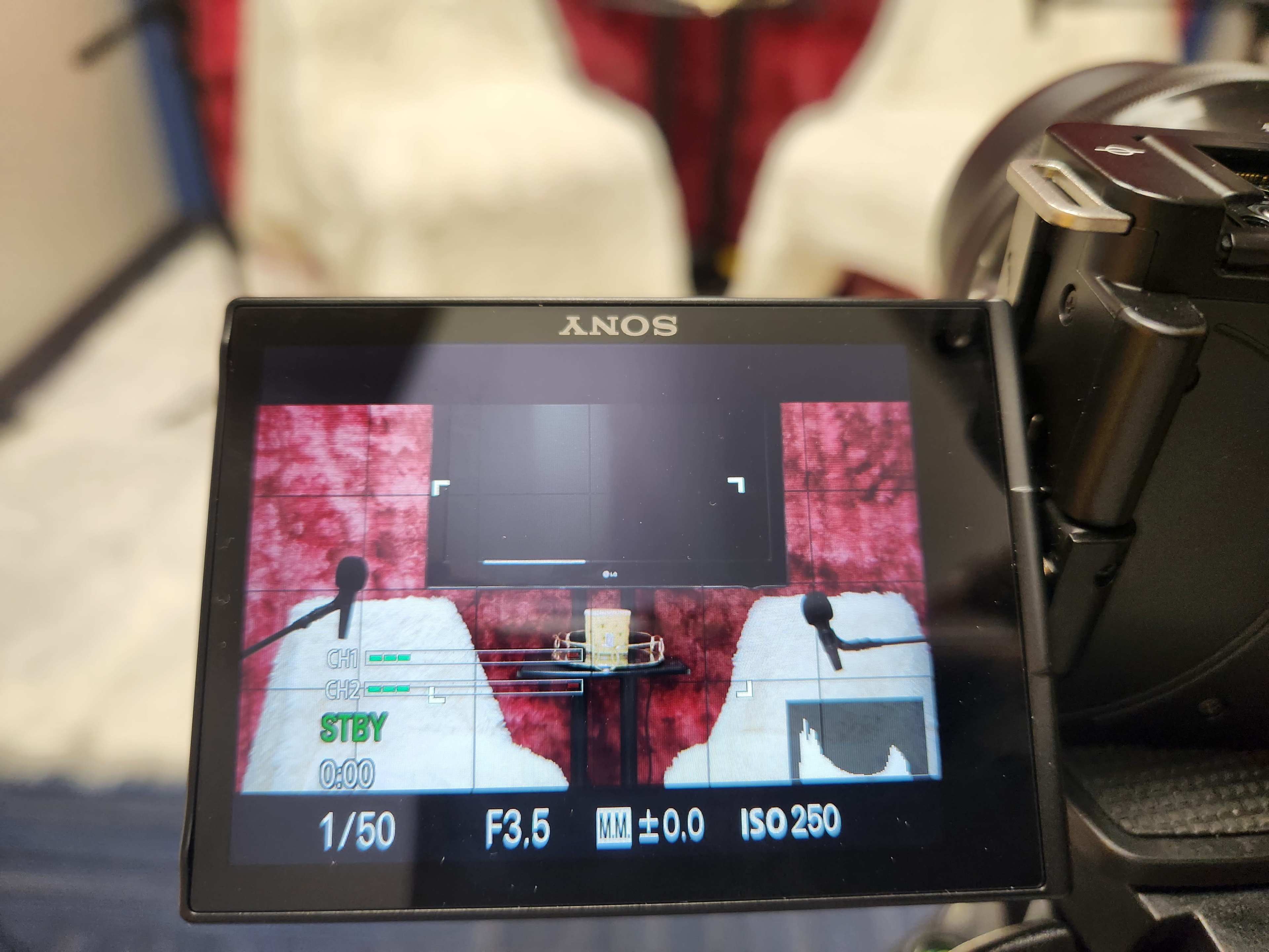 The camera viewfinder shows a setup with two microphones in front of empty white chairs and a screen covered in a red backdrop.