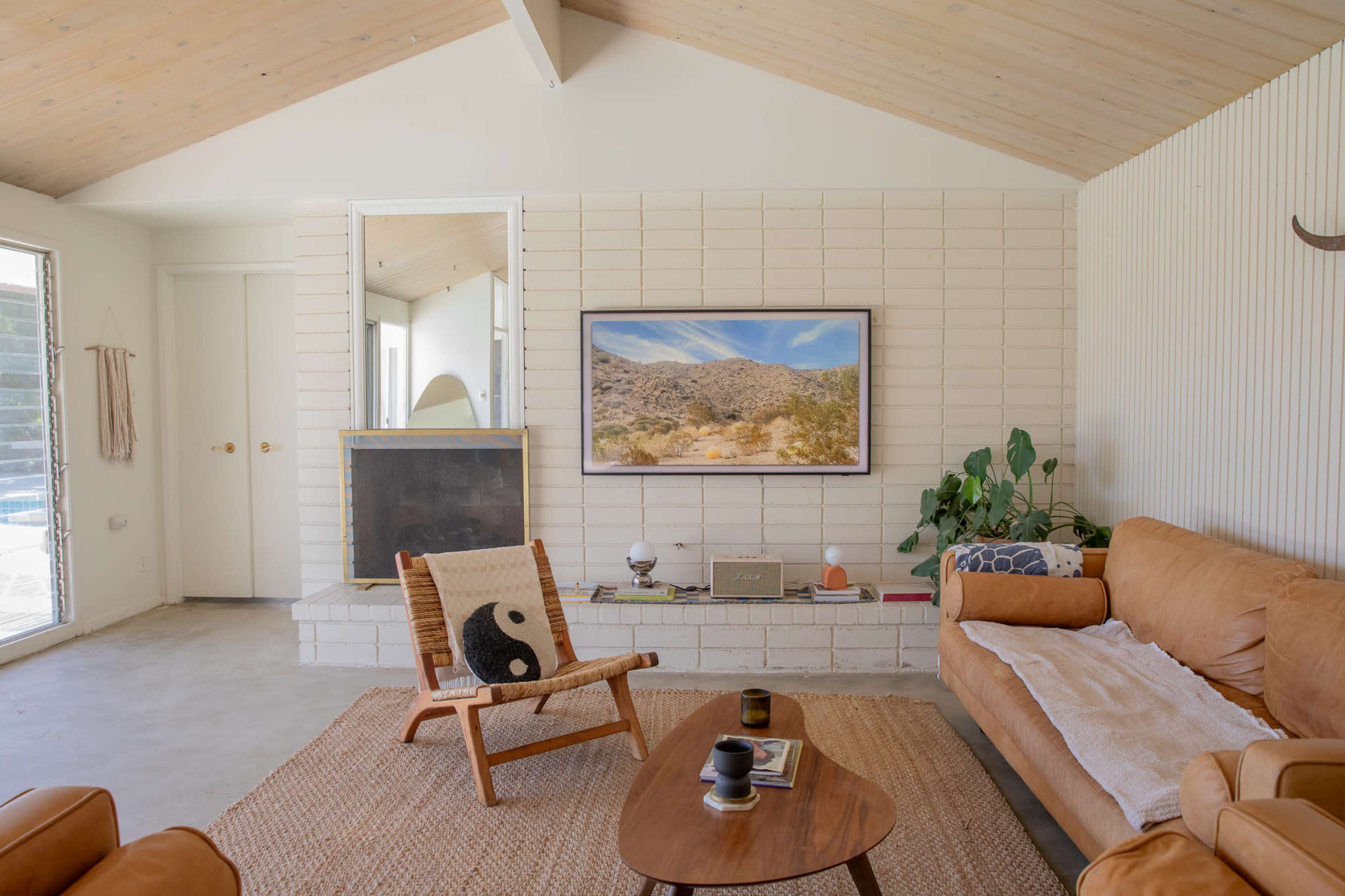 The image shows a modern living room with a tan leather couch, a wooden chair, a coffee table, and a large landscape painting above a white brick fireplace.