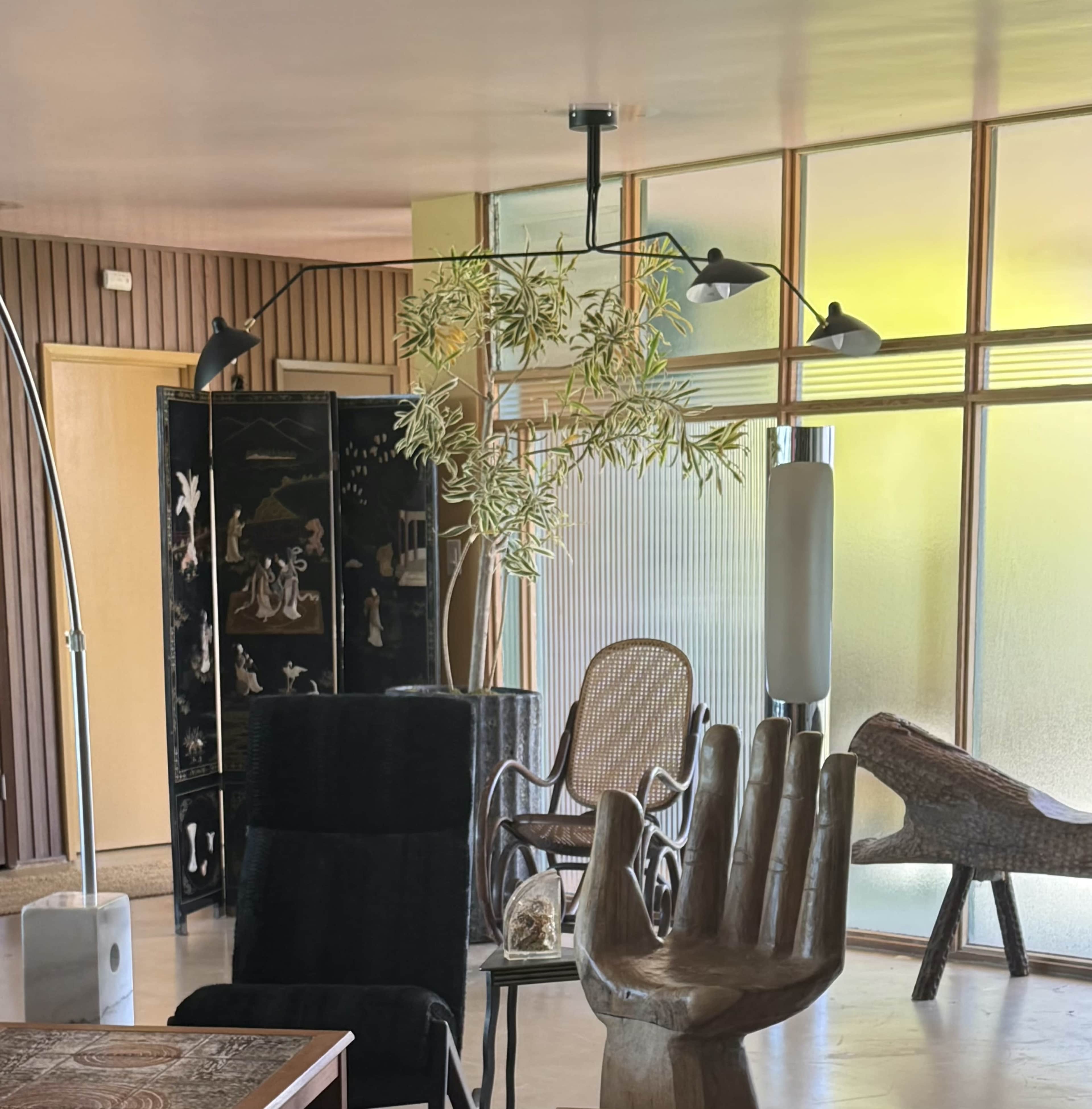 The image shows a modern interior space featuring a mix of furniture, including a wooden hand sculpture, a black chair, and a rattan chair, with large windows allowing natural light to enter.