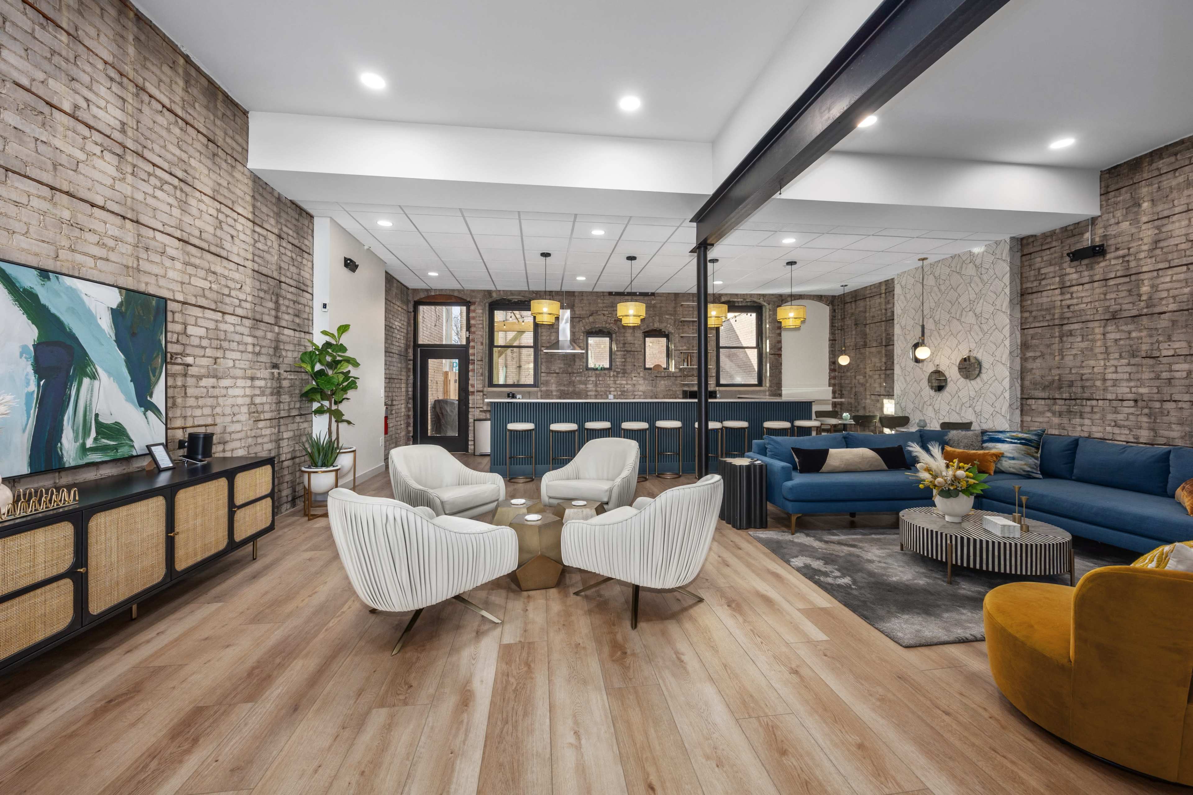 A modern lounge area with a mix of seating options, including two white armchairs, a blue sectional sofa, and a yellow accent chair, surrounded by exposed brick walls and decorative lighting.