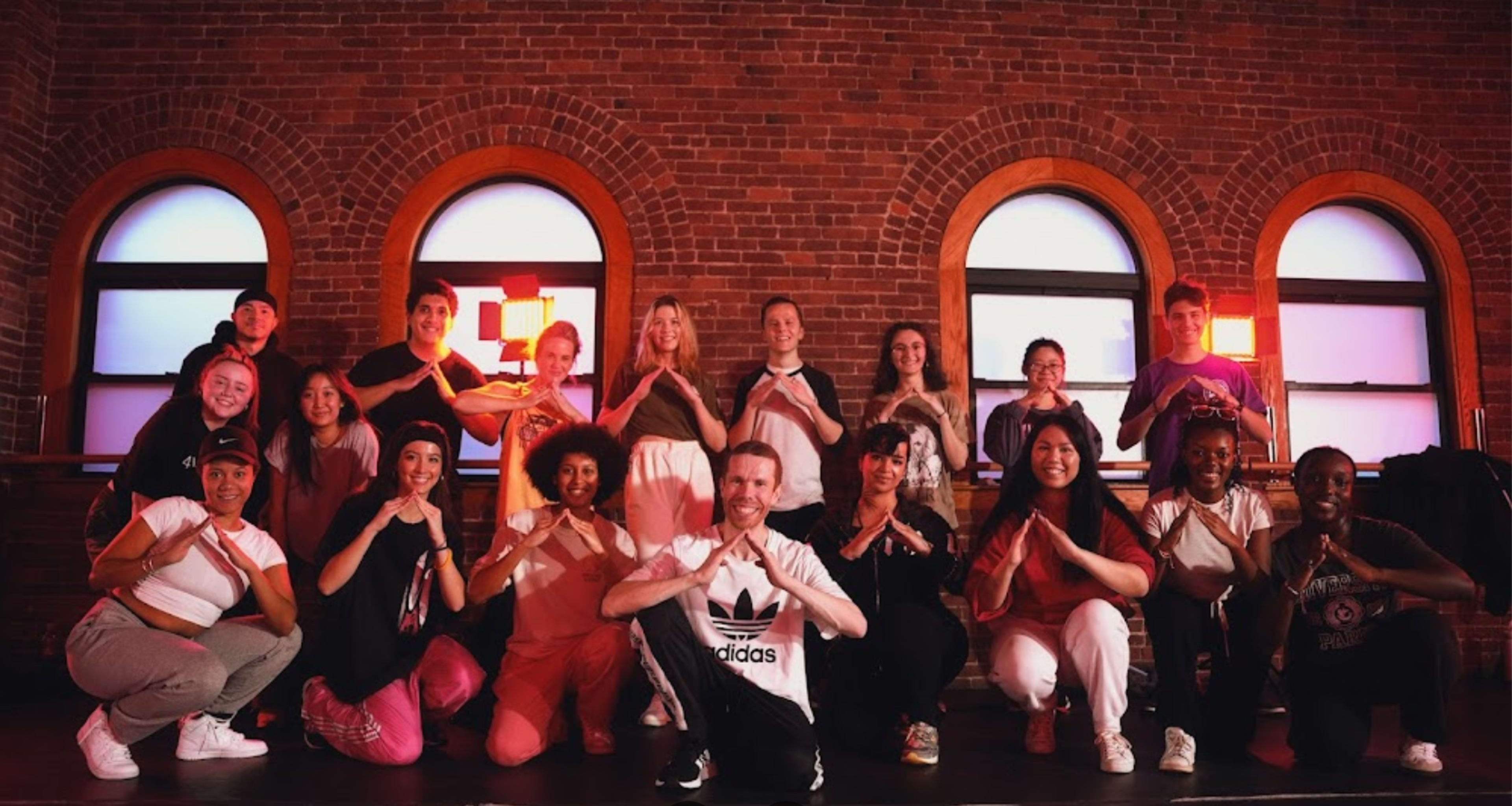 A diverse group of dancers poses together in a well-lit studio with red lamps and brick walls.