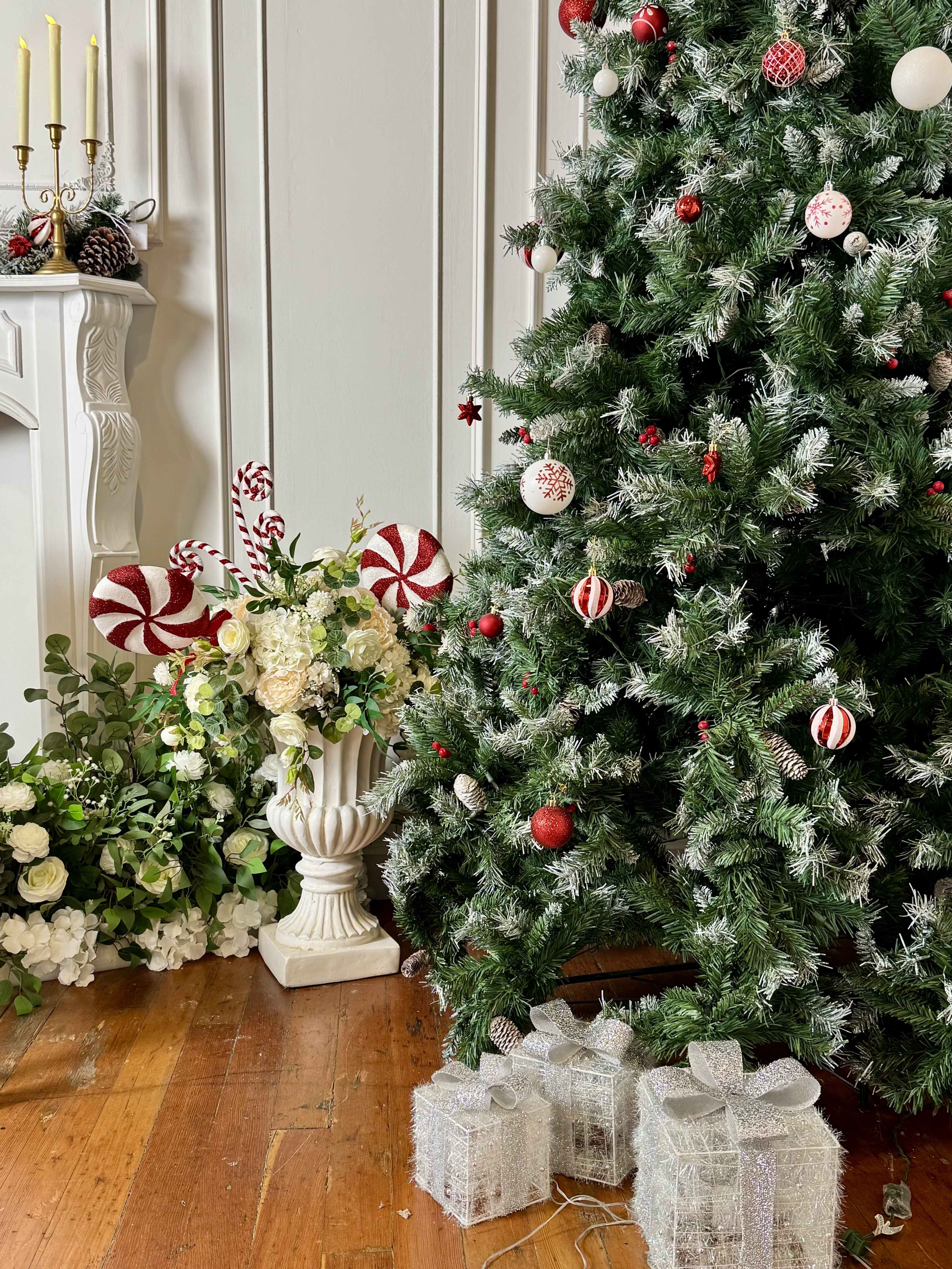 A decorated Christmas tree stands beside a floral arrangement and wrapped gifts on a wooden floor.