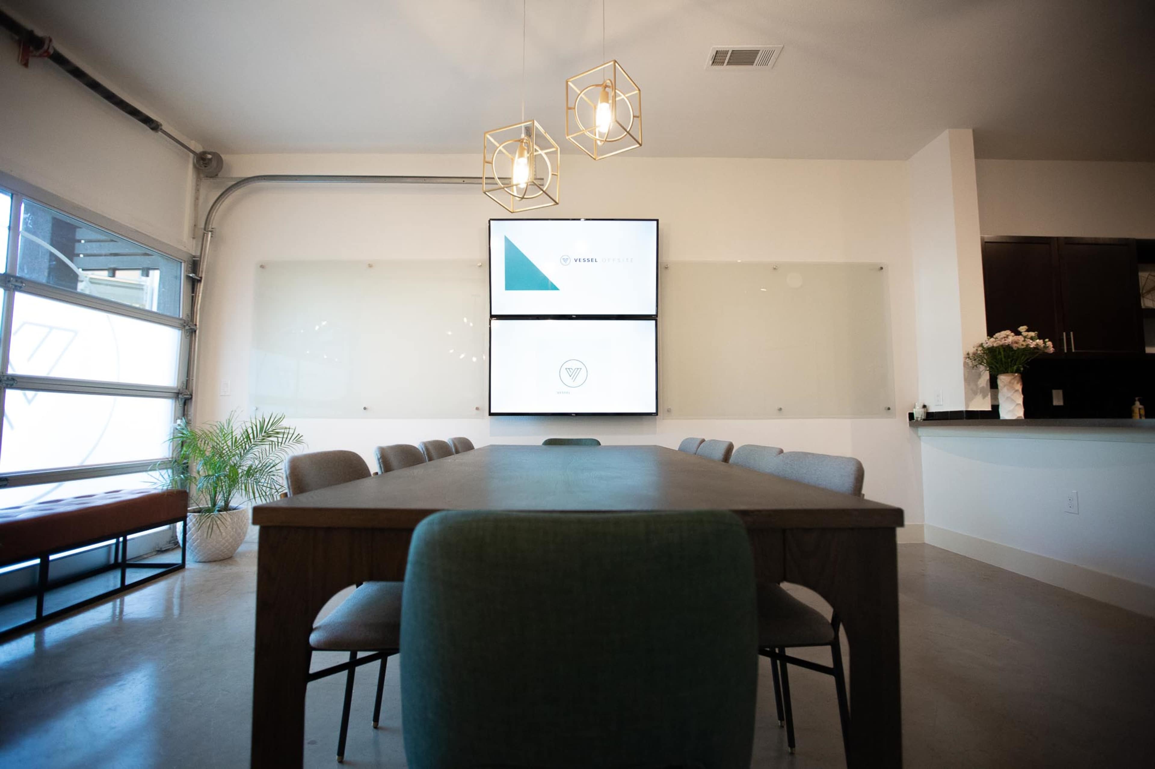 A conference room features a large table surrounded by chairs, with a wall-mounted screen displaying a presentation and decorative lighting above.