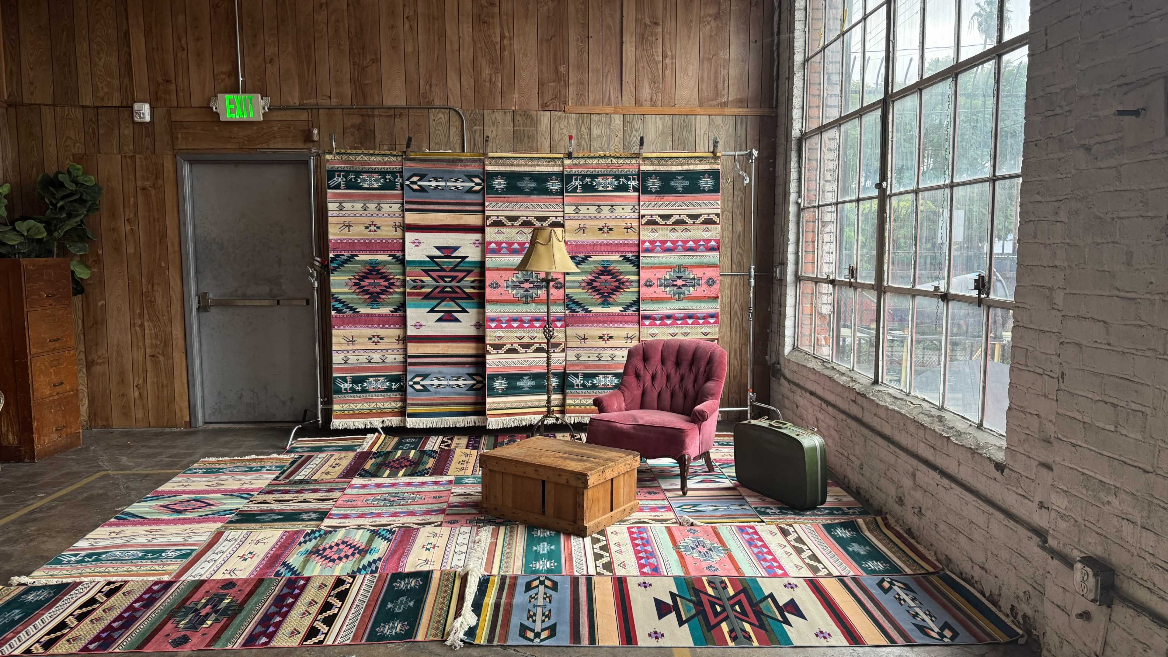 The image shows a cozy interior scene with a pink velvet chair, a wooden chest, and a suitcase situated on a patterned rug, all arranged against a backdrop of colorful woven tapestries and a large window letting in natural light.