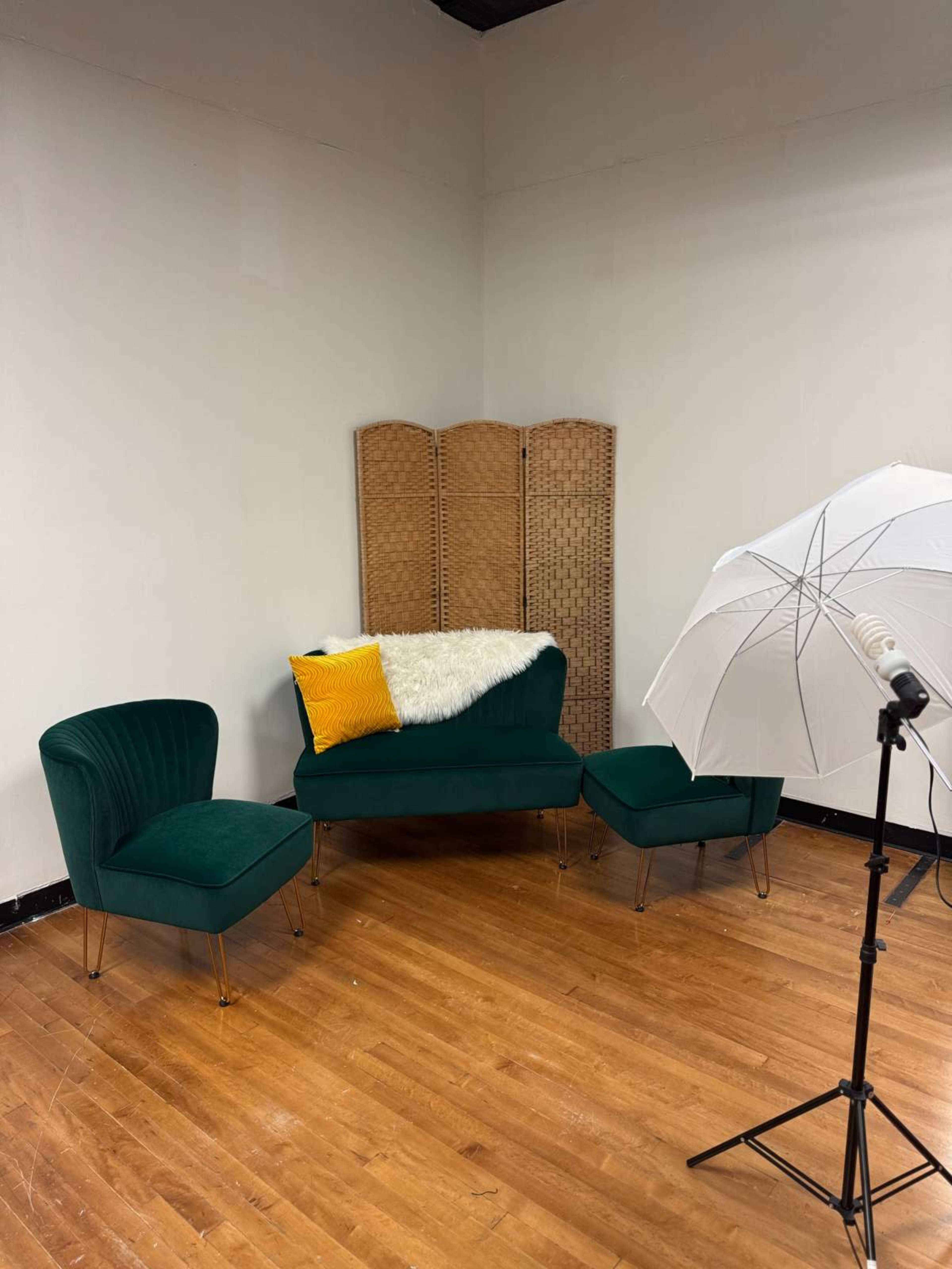 The image shows a small seating area with a green velvet sofa, two matching chairs, a yellow pillow, a white throw blanket, a folding screen in the background, and a lighting umbrella on the side.