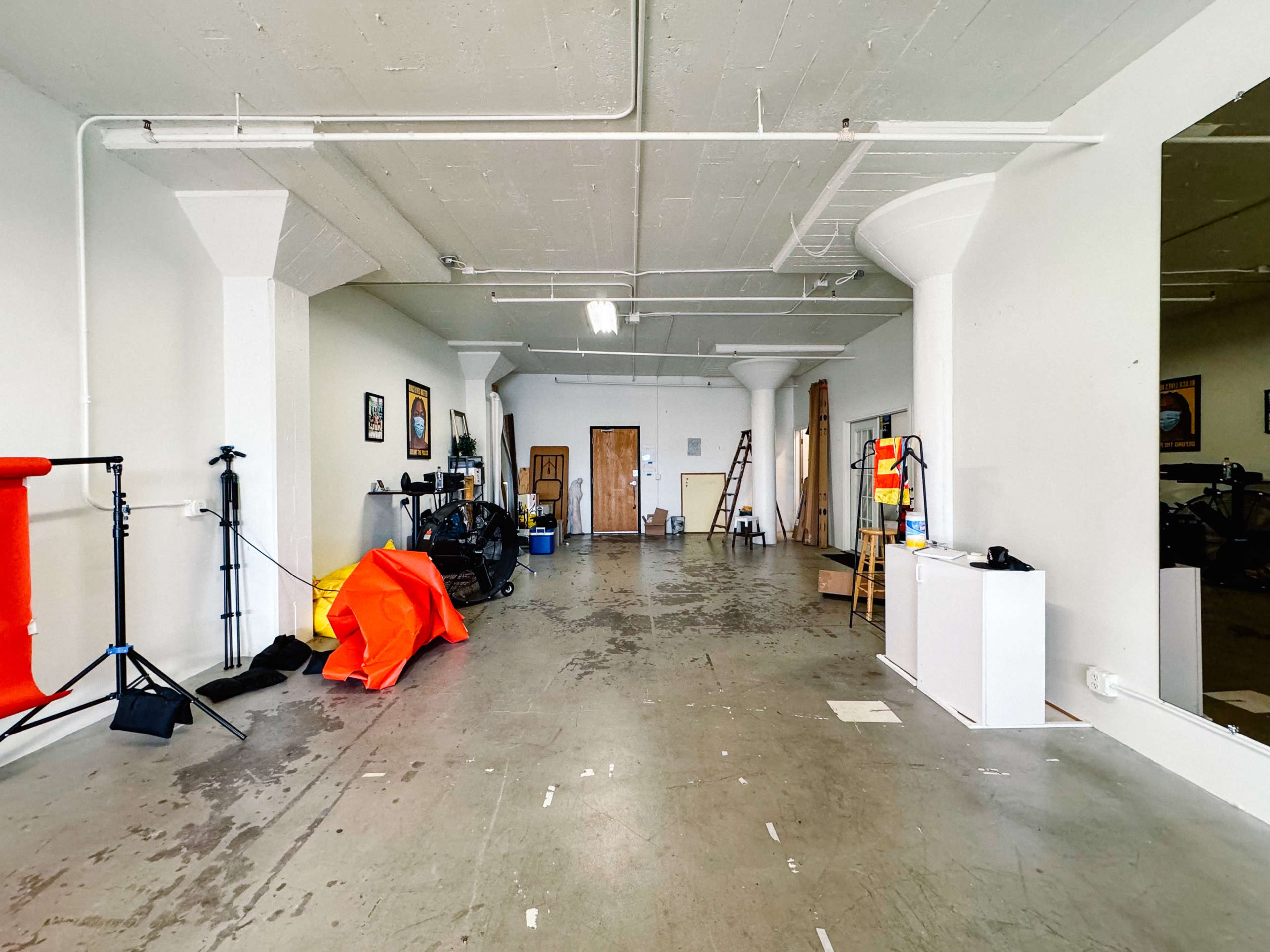 The image shows a spacious, empty room with concrete flooring, white walls, and various equipment and props arranged along one side.